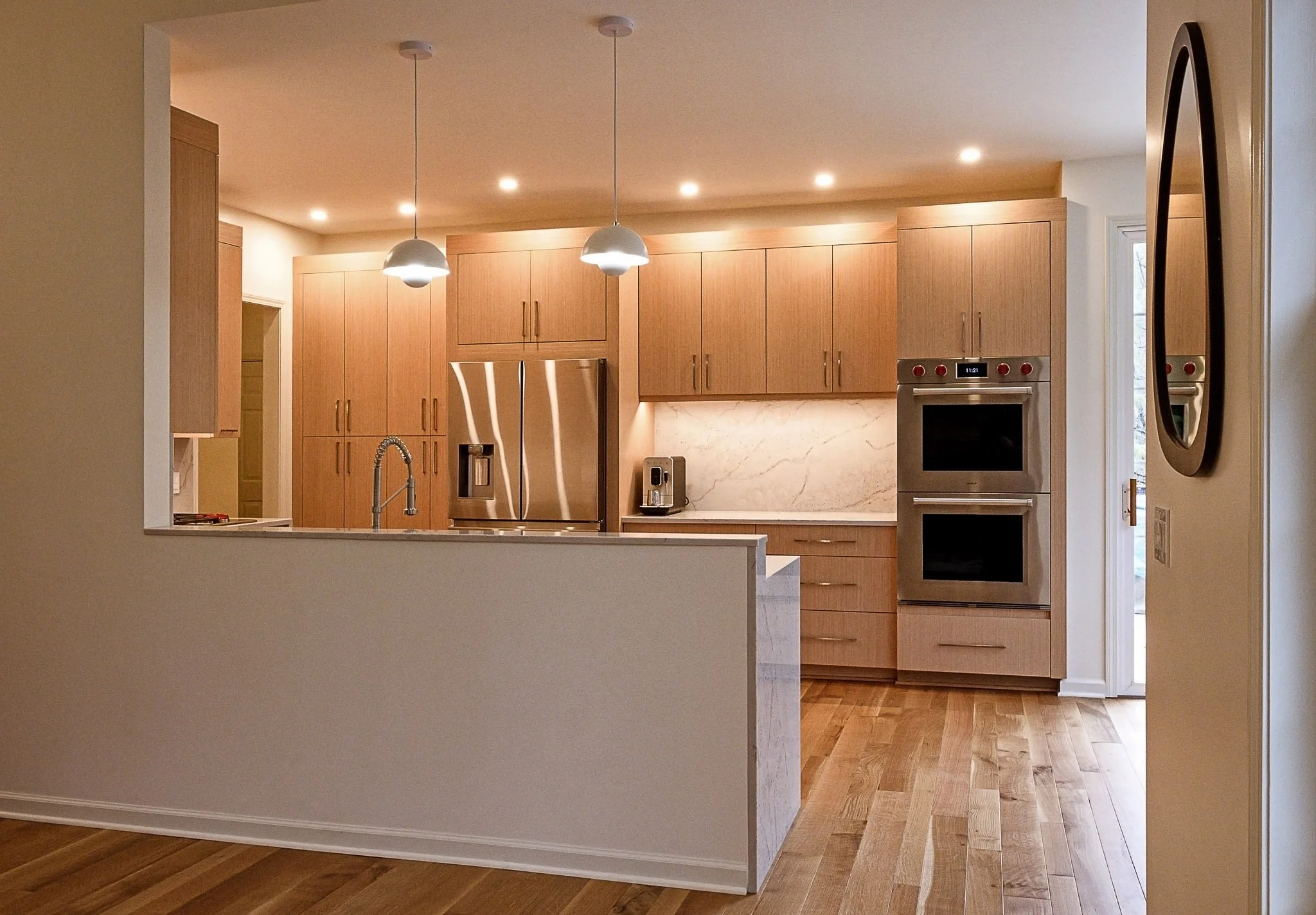 Modern kitchen with wooden cabinets, stainless steel refrigerator, built-in oven, and marble backsplash, wooden flooring, and hanging pendant lights.