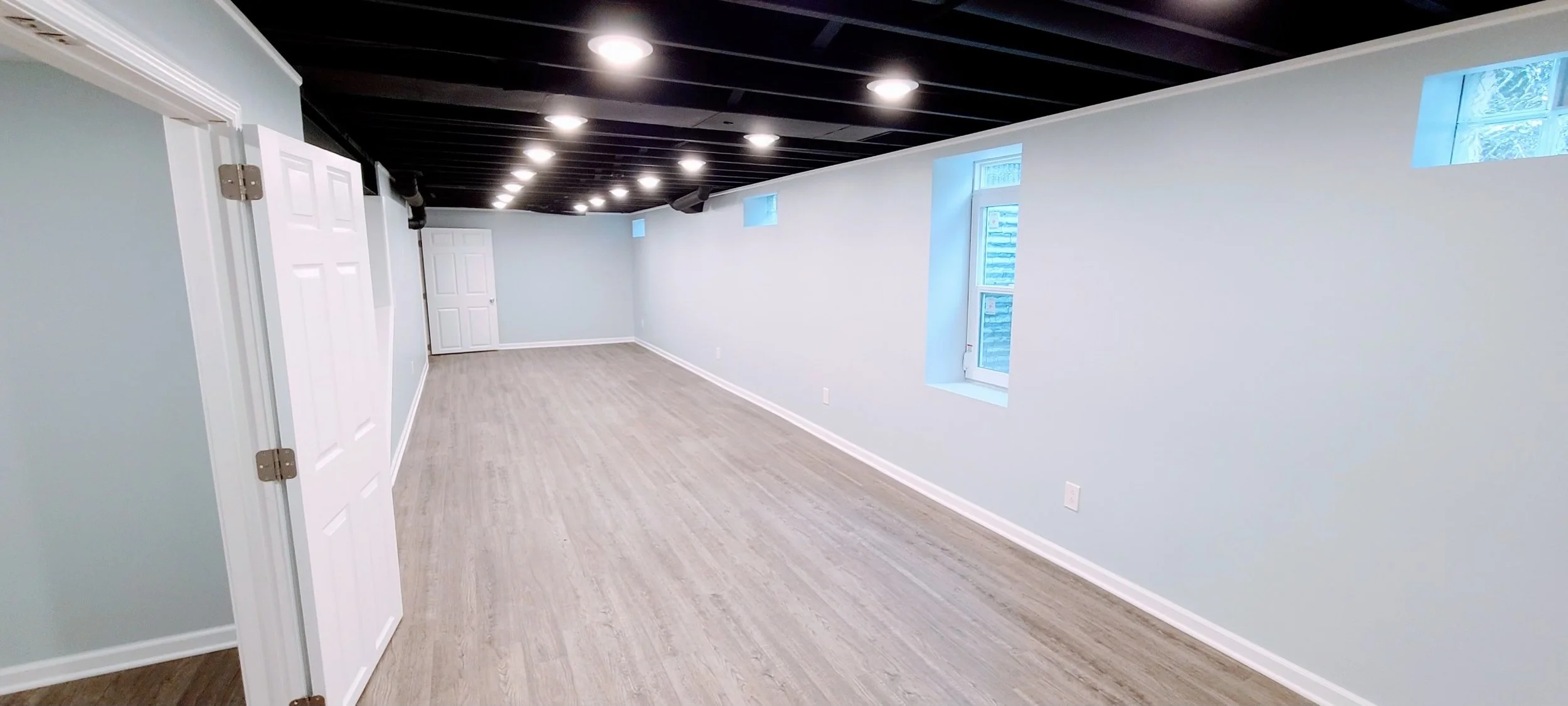 Empty basement room with light-colored wood flooring, white walls, small high windows, and black ceiling with installed lights.