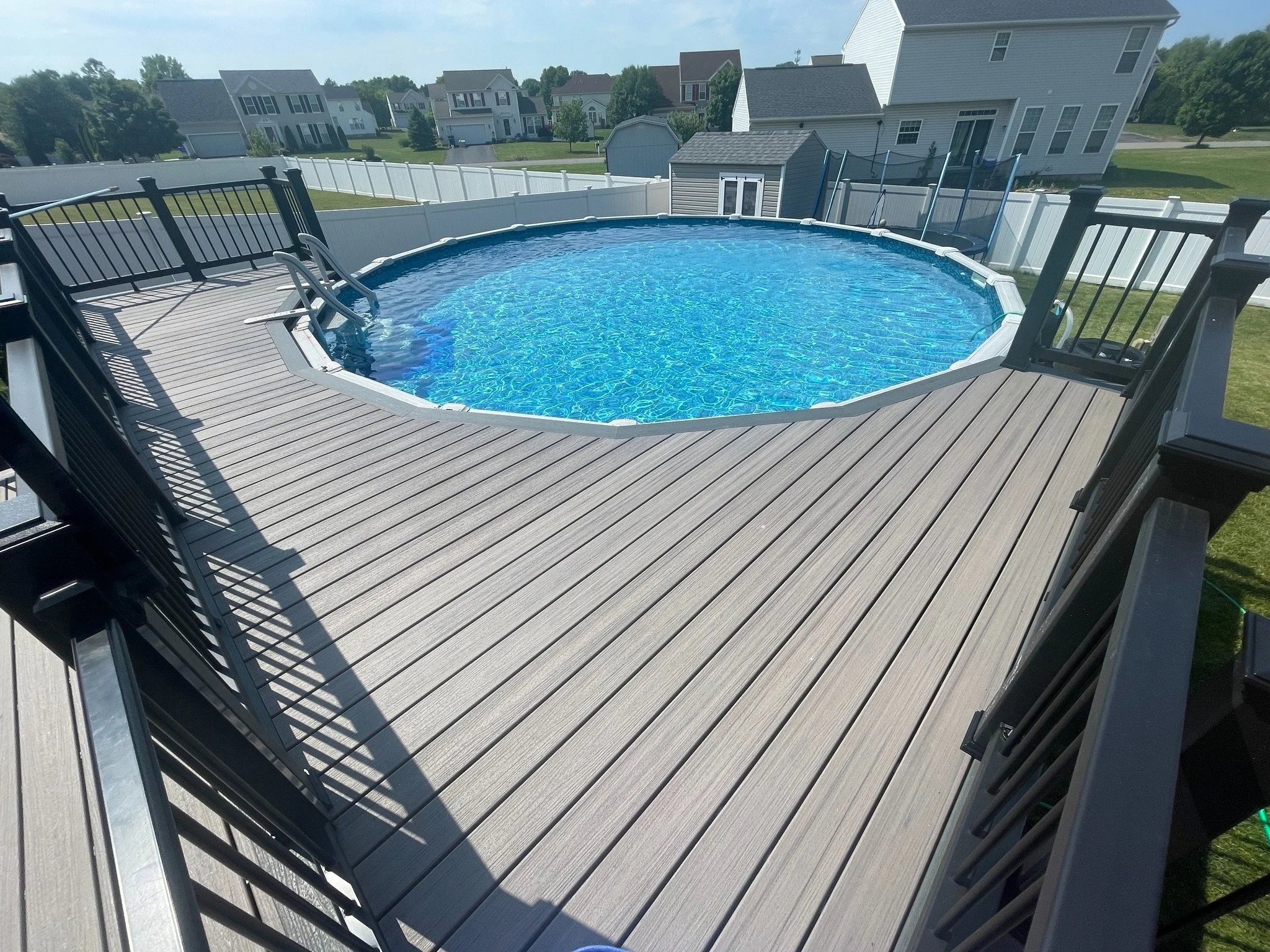 An above-ground swimming pool on a wooden deck in a residential backyard with neighboring houses in the background.