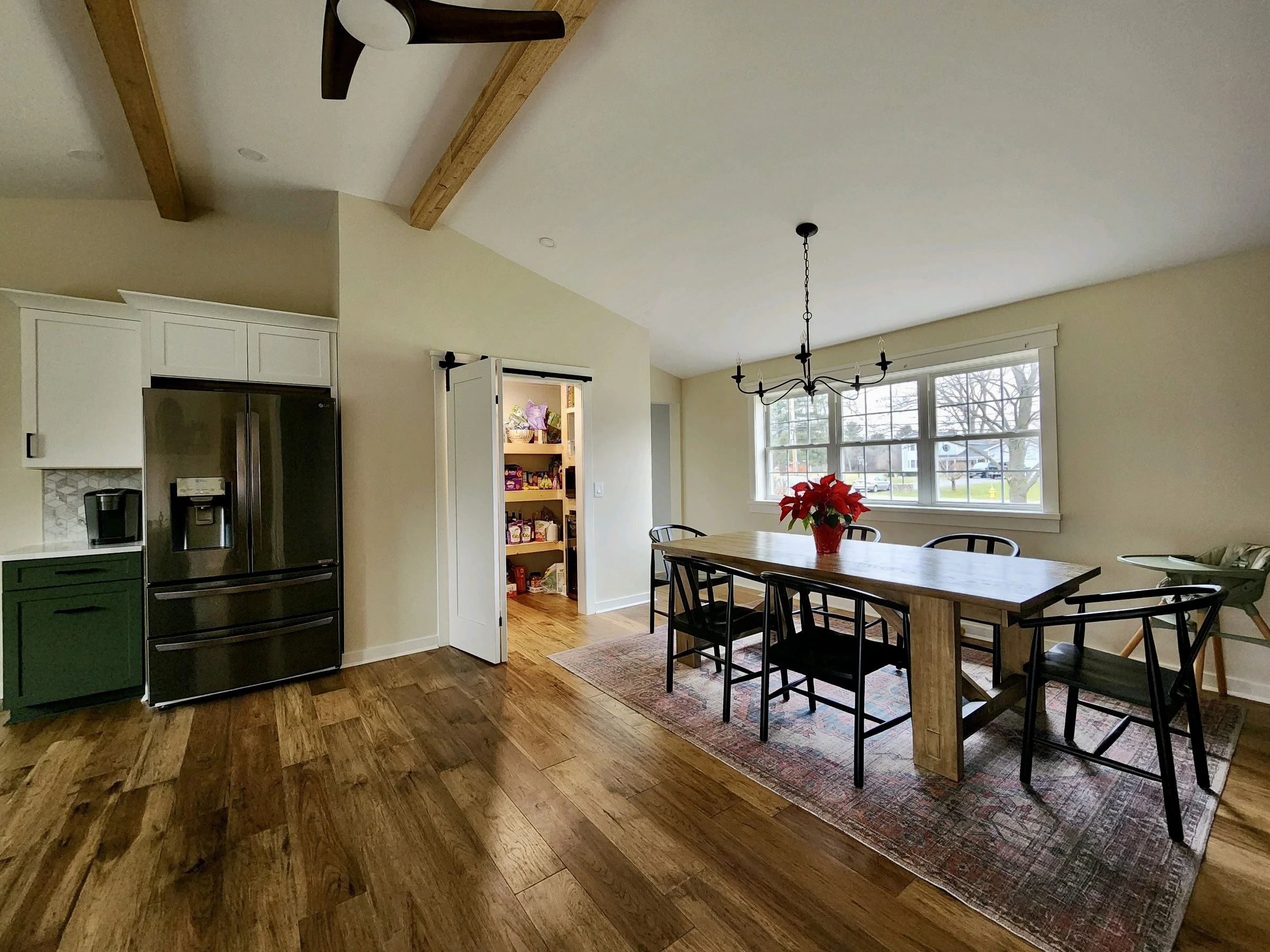 A dining area with a wooden table, six black chairs, and a red poinsettia plant in a red pot. There's a chandelier above the table, three large windows in the background, and a patterned rug under the table. To the left is a kitchen with a stainless 