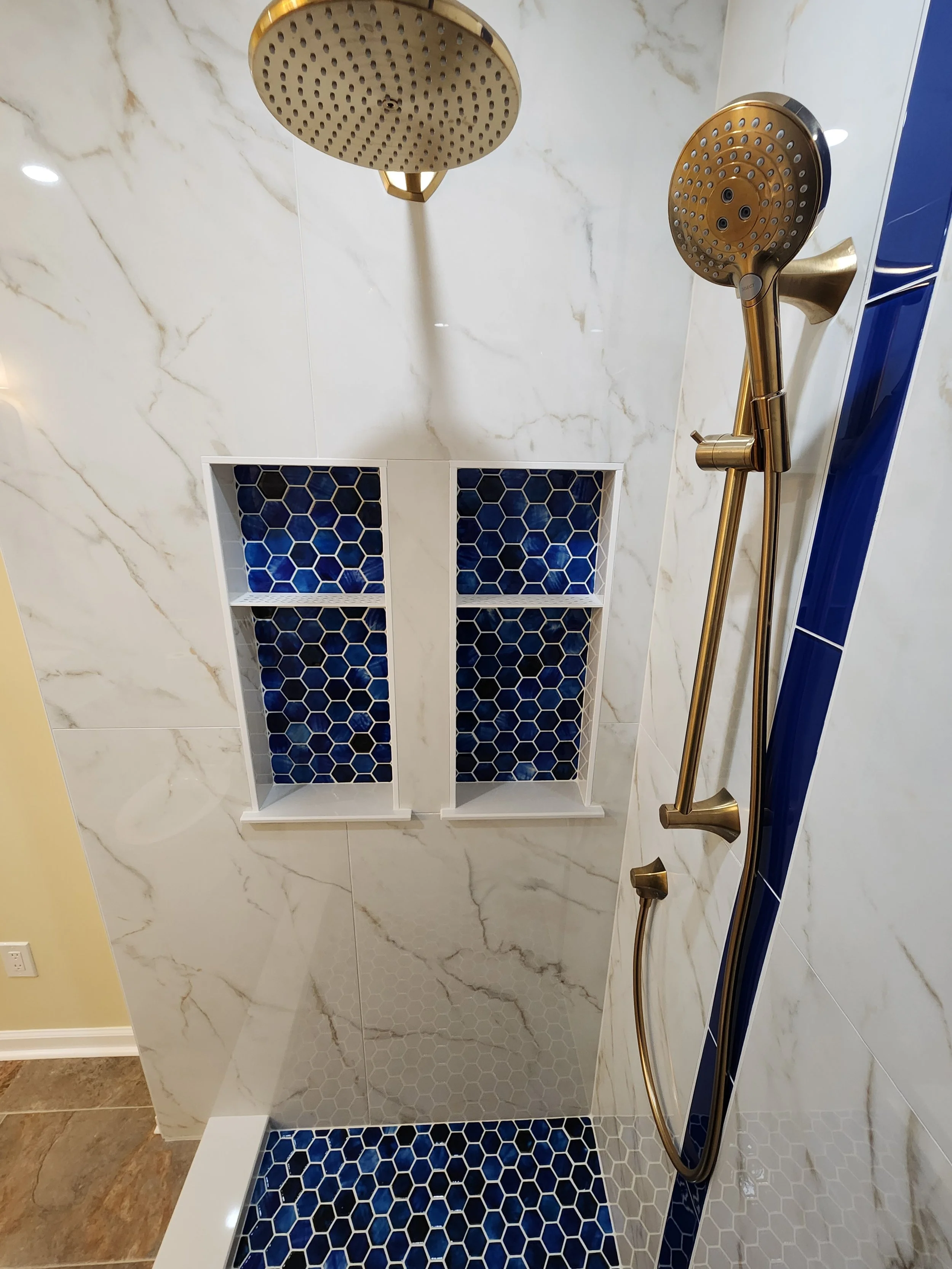 A walk-in shower with marble-patterned walls, two built-in shelves with blue honeycomb tiles, and a brass showerhead and handheld shower.