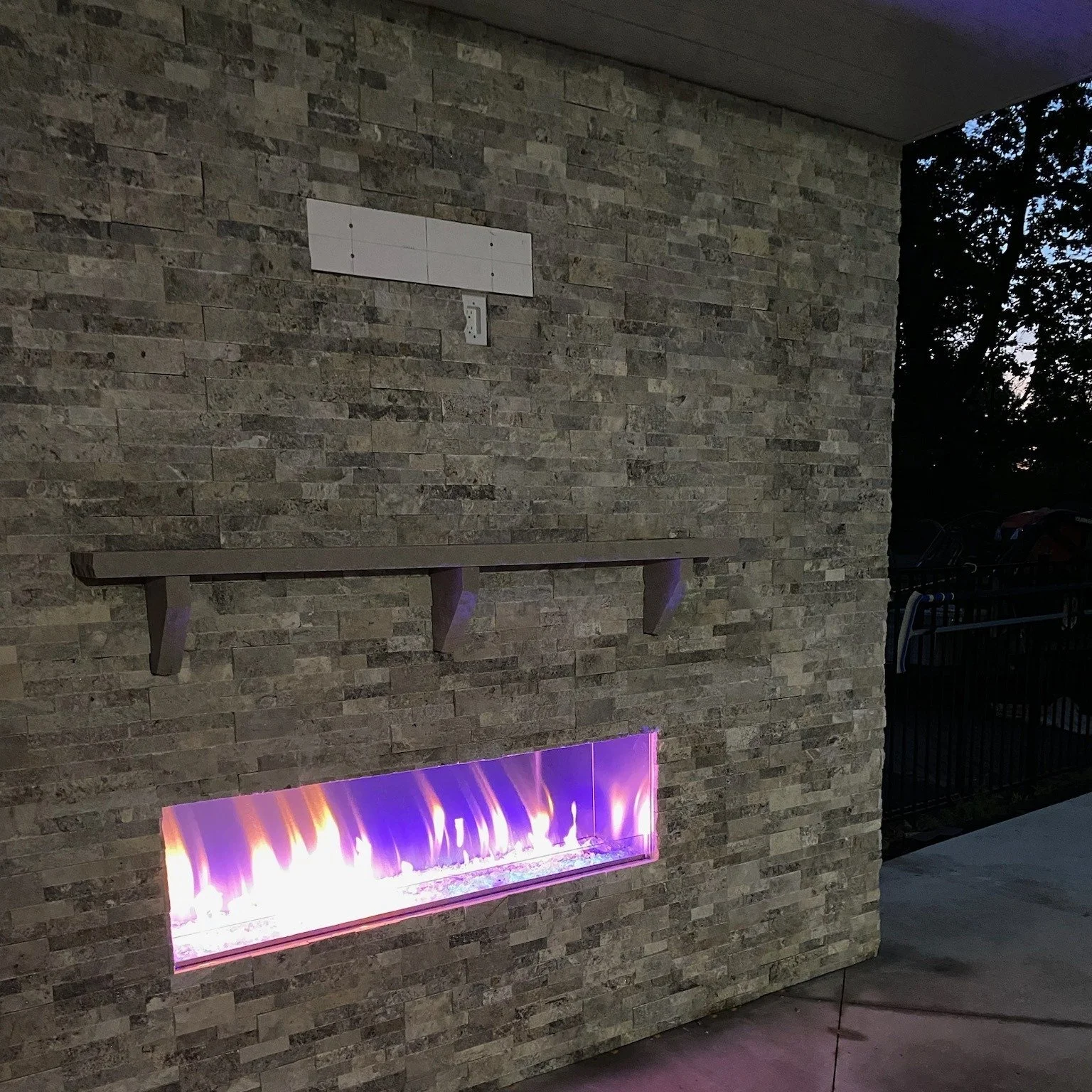 An outdoor installed rectangular gas fireplace with purple and orange flames, built into a stone wall on a patio or porch.