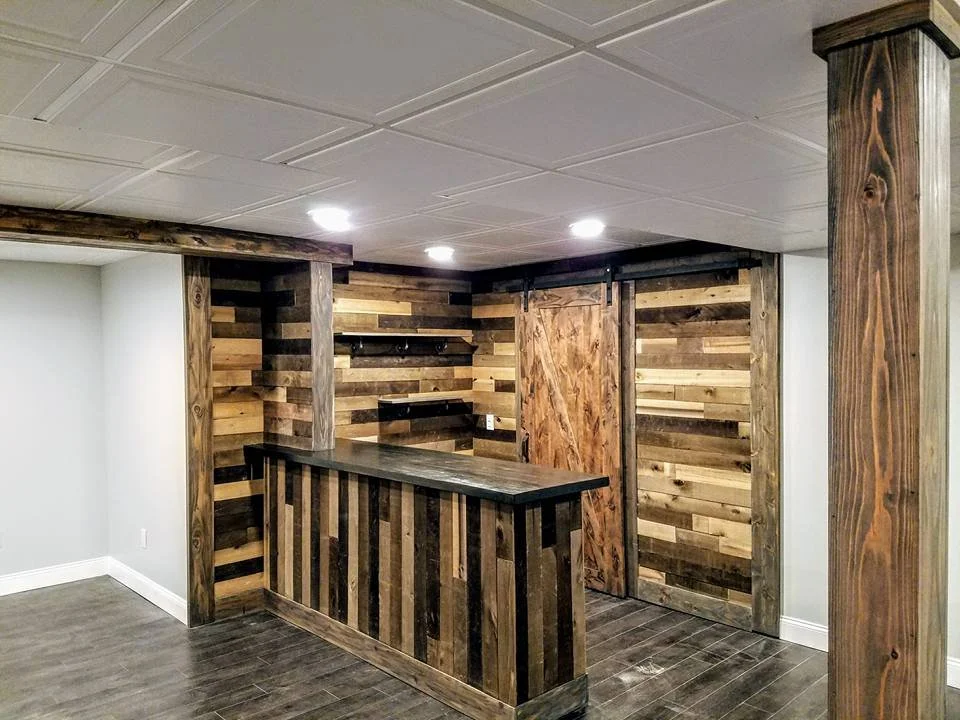 Interior of a room with a wooden bar counter and wood-paneled walls, ceiling lights, and a rustic door.