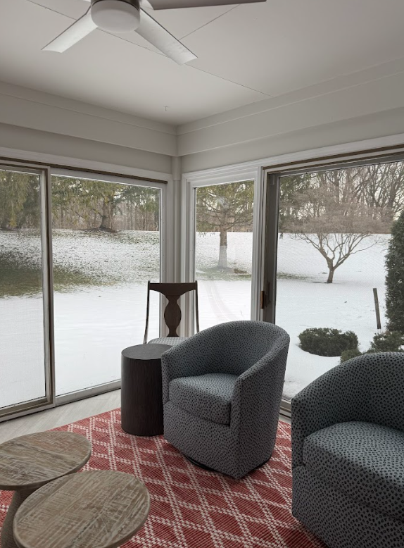Sunroom with large windows showing snowy landscape and trees outside, containing two patterned armchairs, a side table, and a ceiling fan.