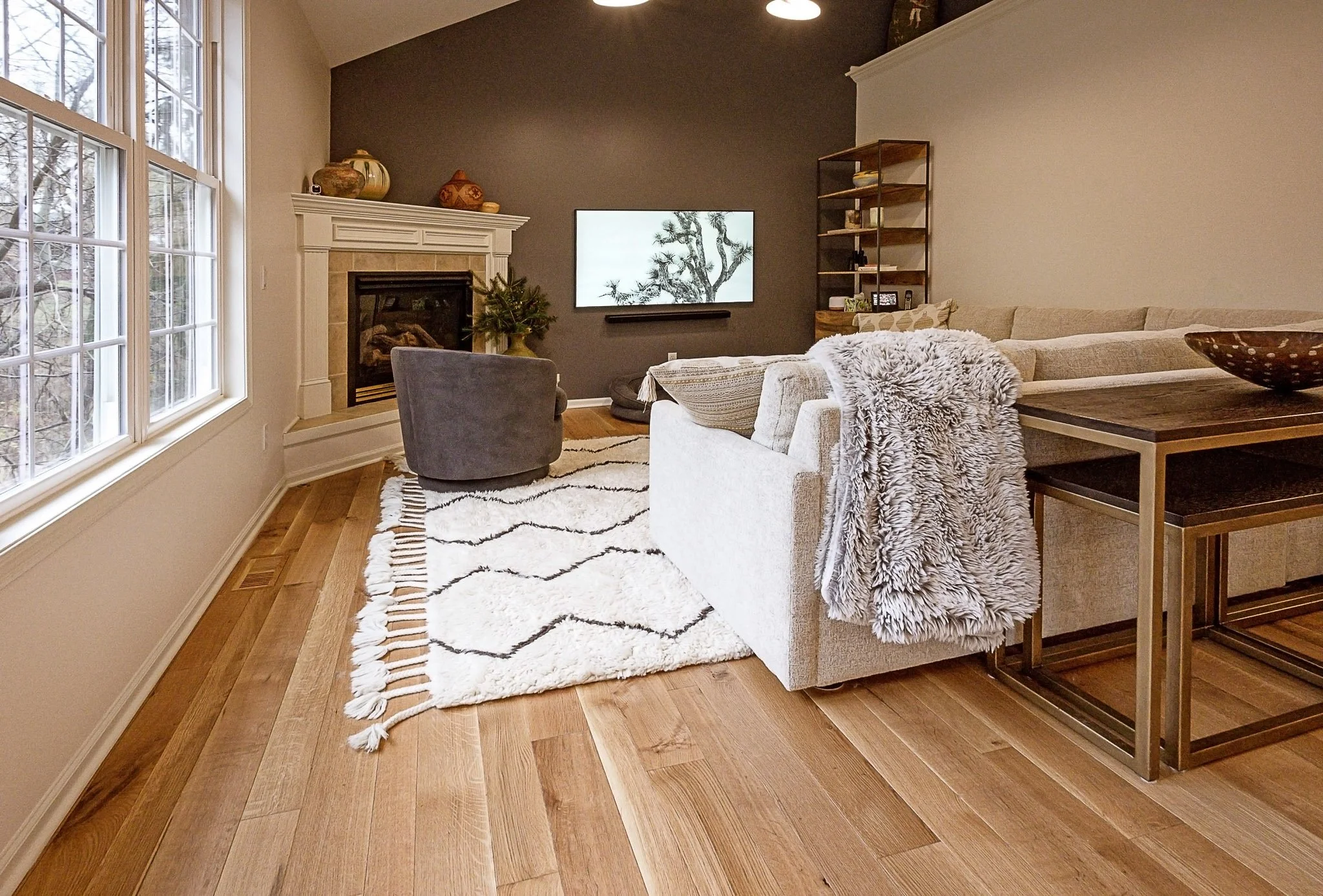 Living room with large windows, a fireplace, a wall-mounted TV, a beige sectional sofa with a fluffy throw, a gray accent chair, a striped rug, a wooden side table, and a built-in bookshelf.