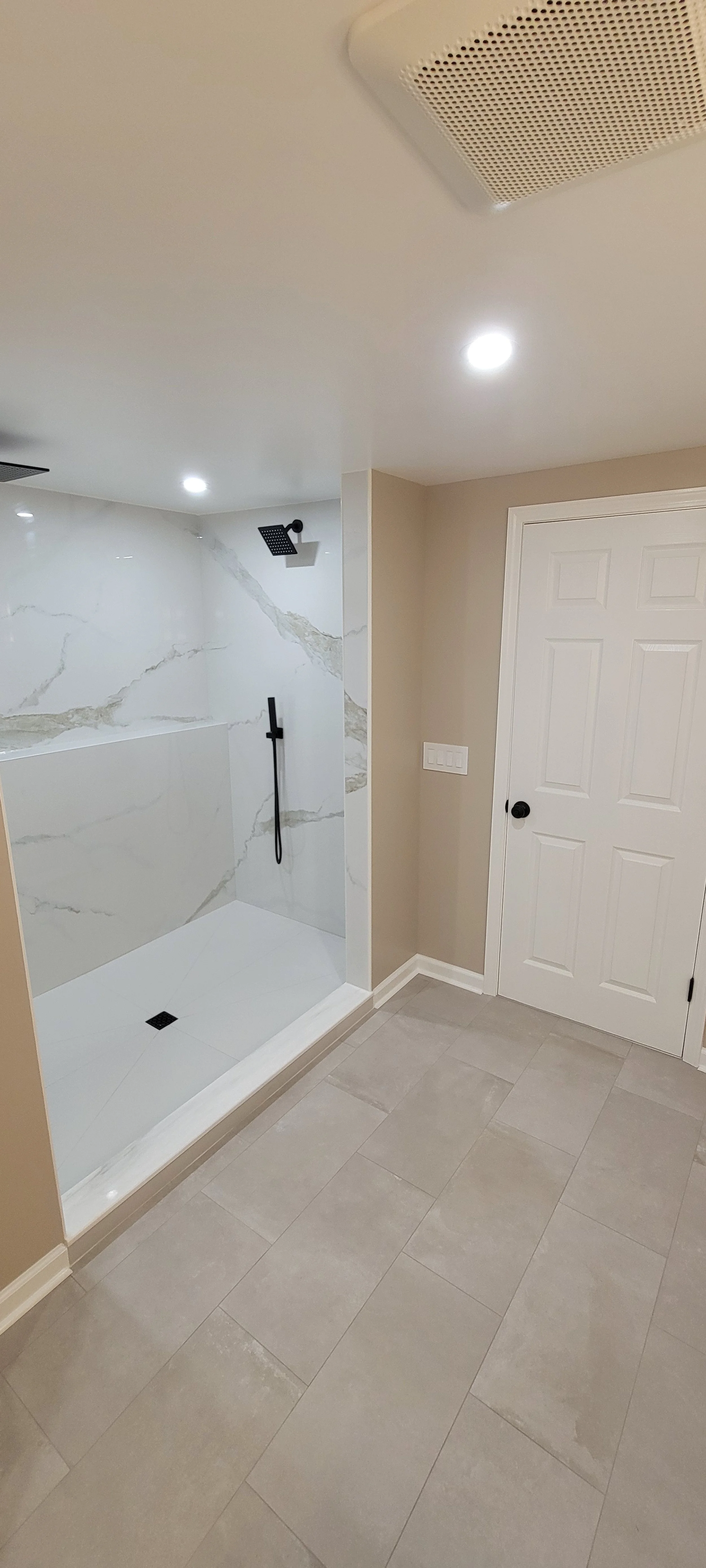 Bathroom with beige tiled floor, white door with black handle, and walk-in shower with white marble walls, black showerhead, and beige fixtures.