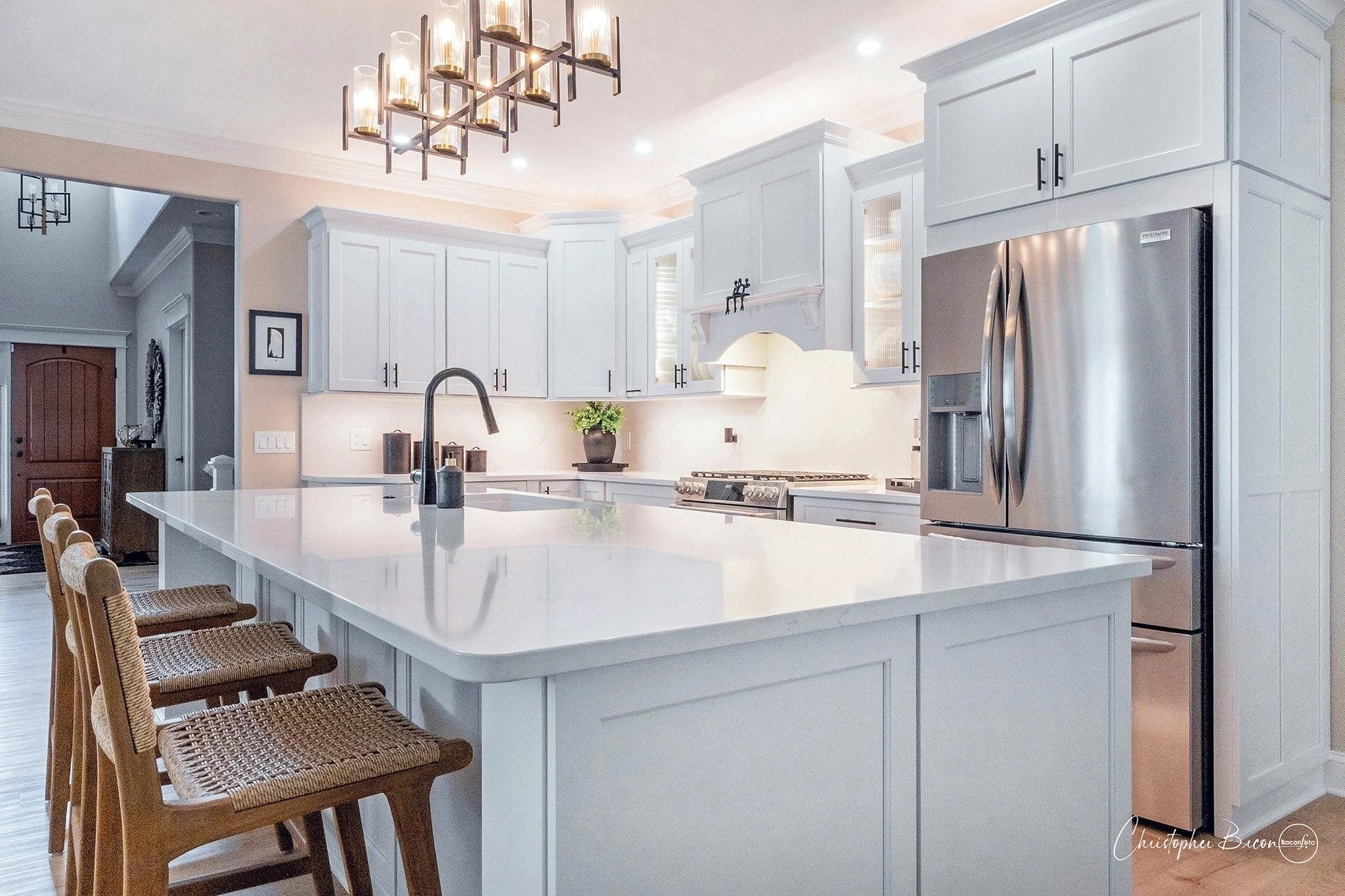 Modern kitchen with white cabinets, a large island with a white countertop, a stainless steel refrigerator, a black faucet, and woven bar stools.