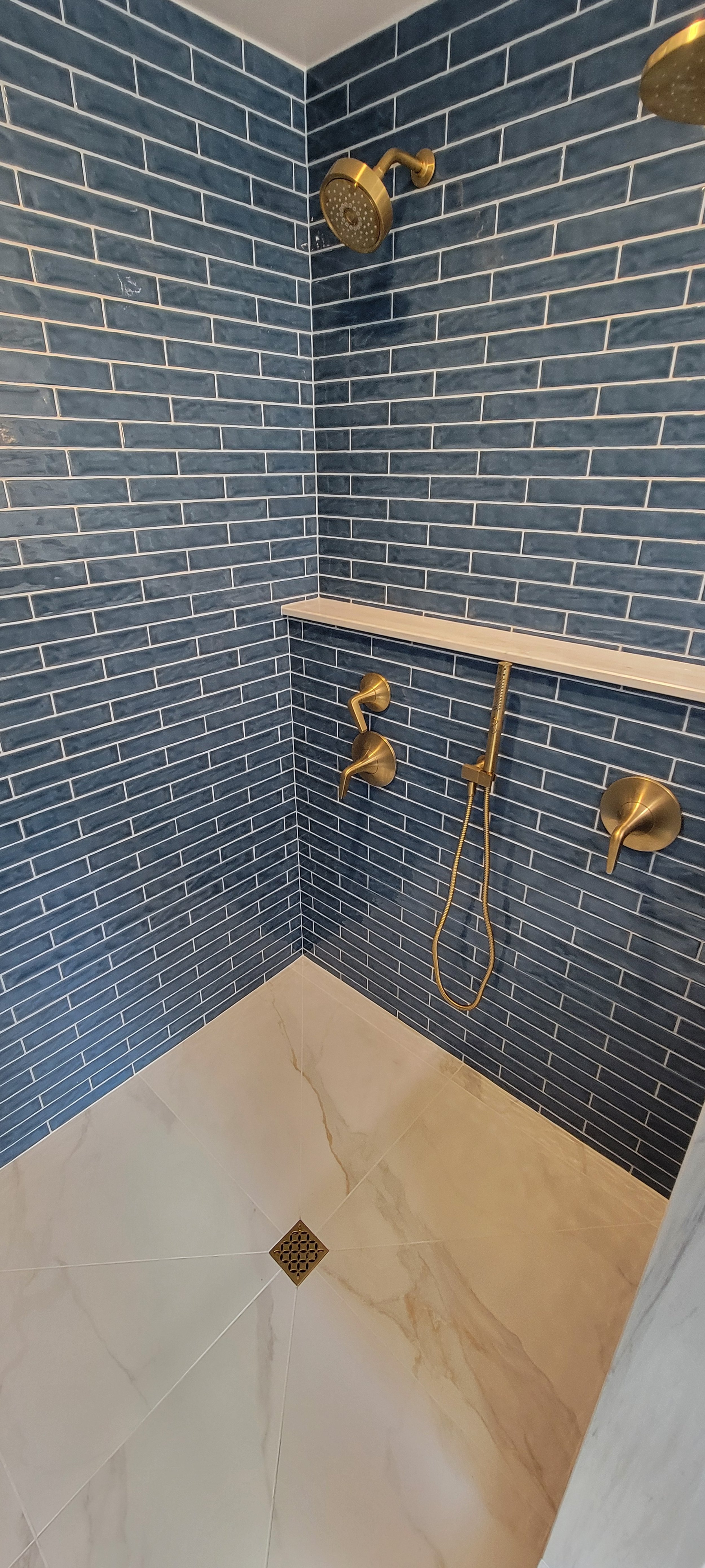 A modern walk-in shower with blue subway tile walls, gold fixtures, a hand-held showerhead, and a small built-in shelf.
