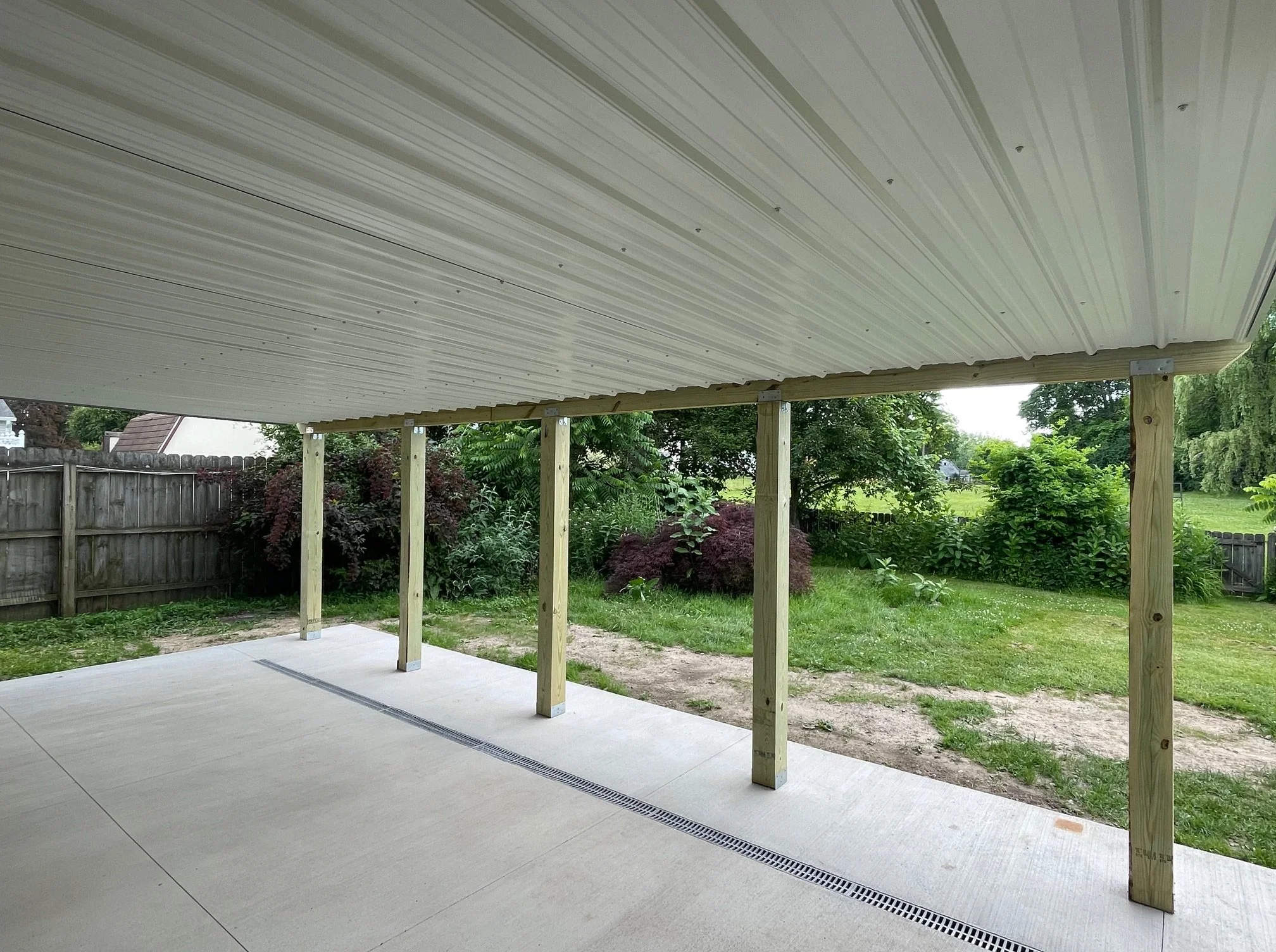 View of a backyard patio under construction with new wooden support beams and a concrete floor, surrounded by a green grassy area and trees.