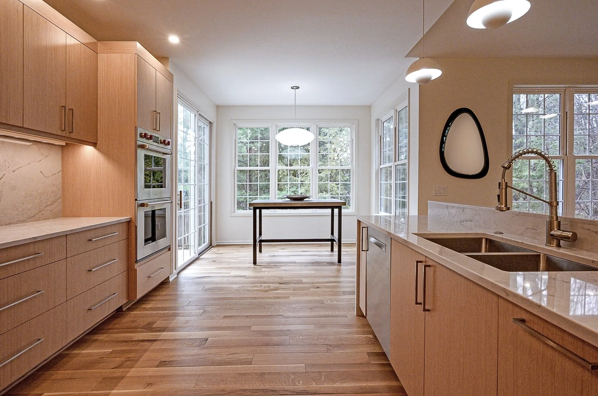 Bright, modern kitchen with wooden cabinets, marble countertops, a double sink with a gold faucet, hardwood flooring, and large windows overlooking trees. There is a small table near the windows and contemporary pendant lighting.
