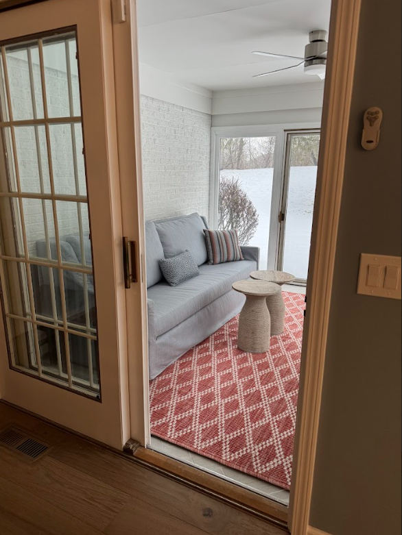 View through a door shows a sunlit sitting area with a gray sofa, two striped cushions, and two textured cushions, a patterned red and white rug, and a small beige side table. Large window/door reveals a snow-covered outdoor area with a bush.