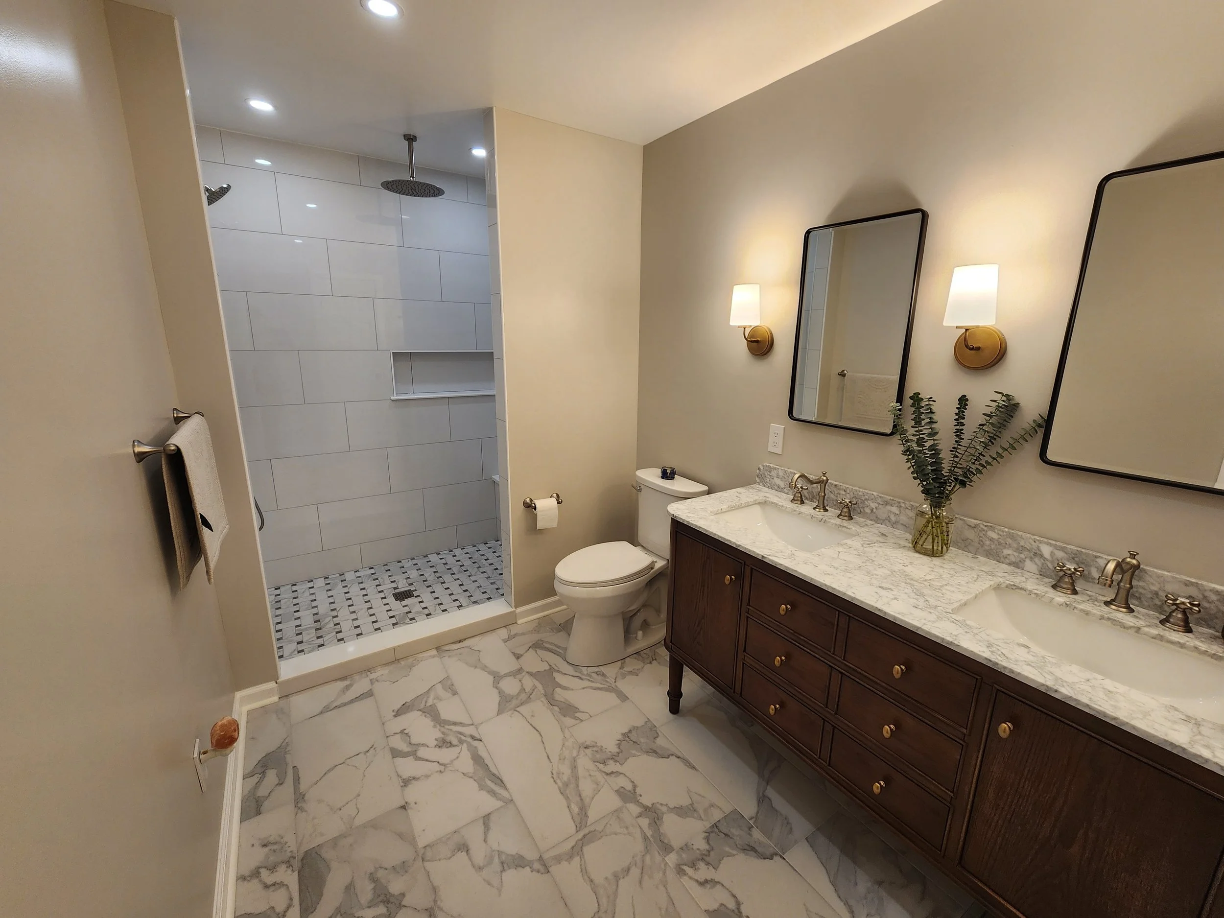 Modern bathroom with a marble floor, double vanity with a marble countertop, two mirrors, and two wall-mounted lights. There is a toilet and a walk-in shower with tiled walls and a rain showerhead.