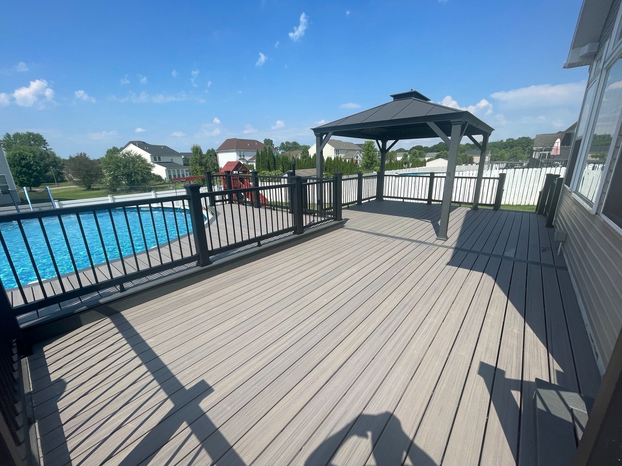A large outdoor wooden deck with black metal railing, a gazebo, overlooking a swimming pool, neighboring houses, green trees, and a blue sky with clouds.