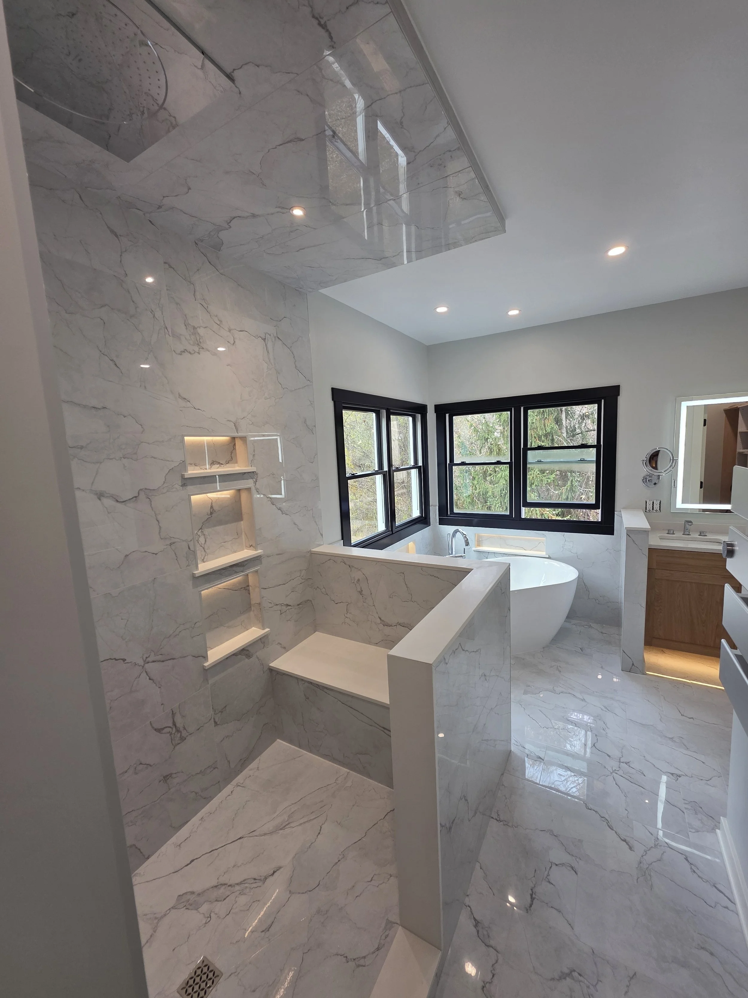 Modern bathroom with marble walls and floors, a freestanding bathtub near windows, a walk-in shower with built-in shelves, a wooden vanity with lighting, and black window frames.