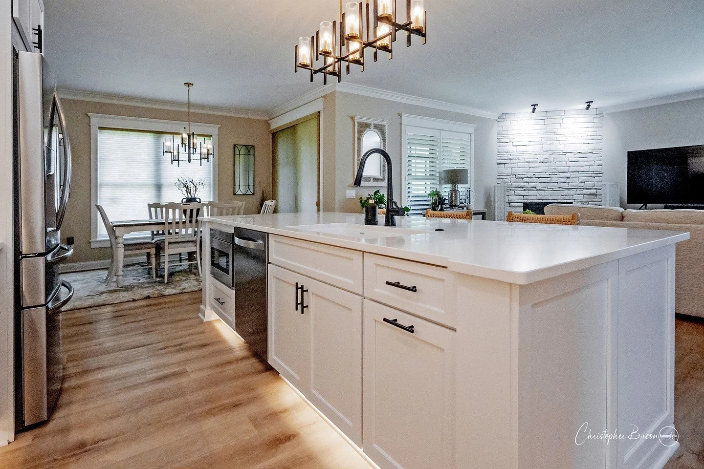An open-concept kitchen and living room with a large white island, black hardware, a stainless-steel refrigerator, and a black oven. There's a dining area with a window, a chandelier, a white dining table, and chairs. The living room features a stone accent wall with lighting, a large television, and a comfortable beige sectional sofa.