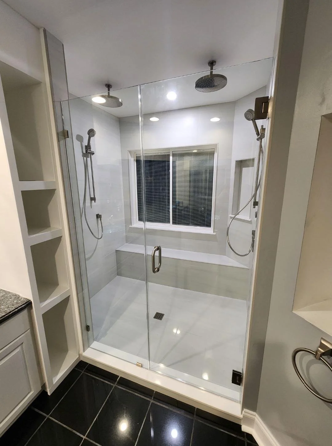 Modern bathroom with a glass shower enclosure, two showerheads, a window, and black tiled flooring.