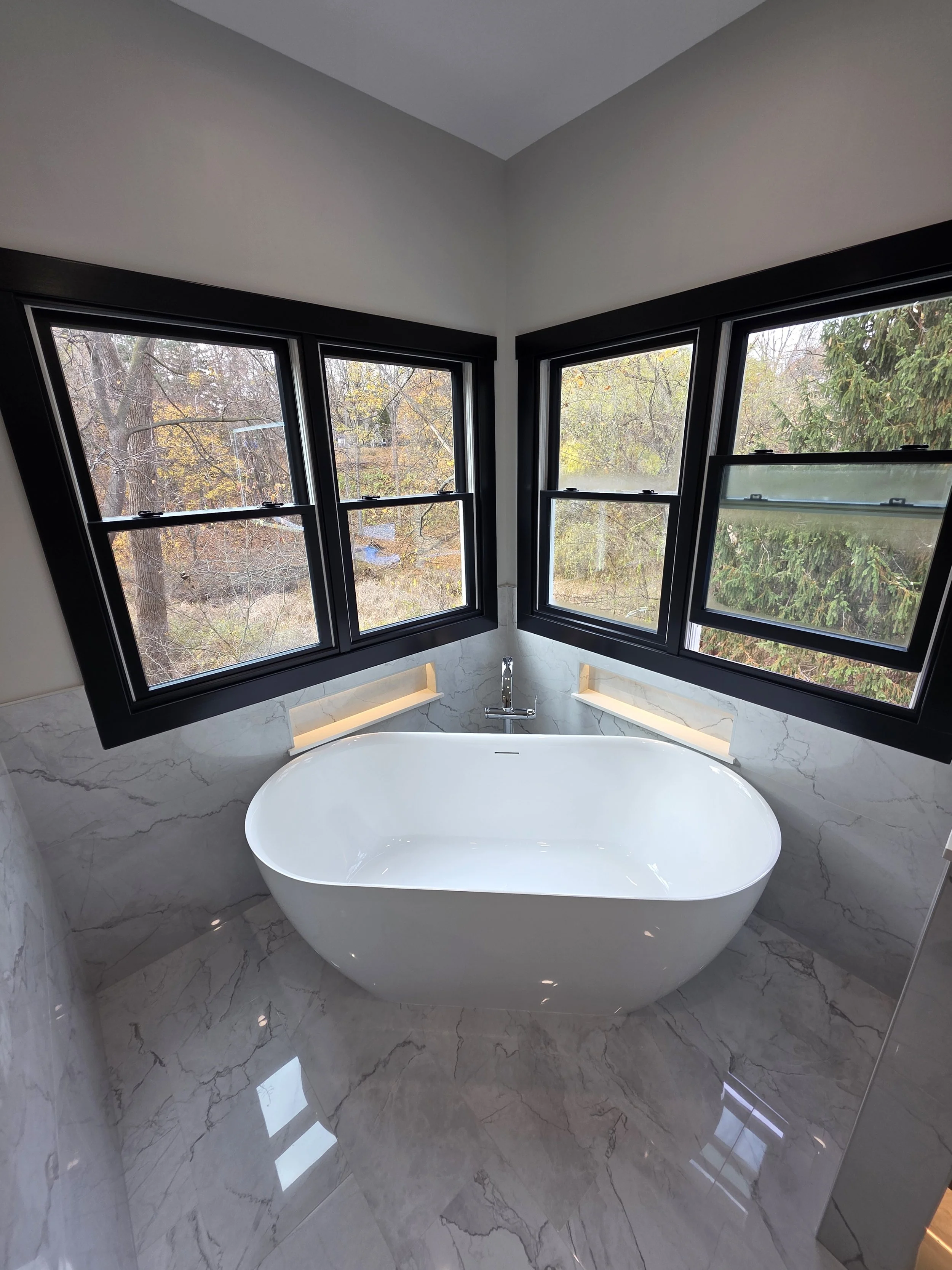 Modern bathroom with a free-standing bathtub beneath four black-framed windows, marble walls, and a view of trees outside.