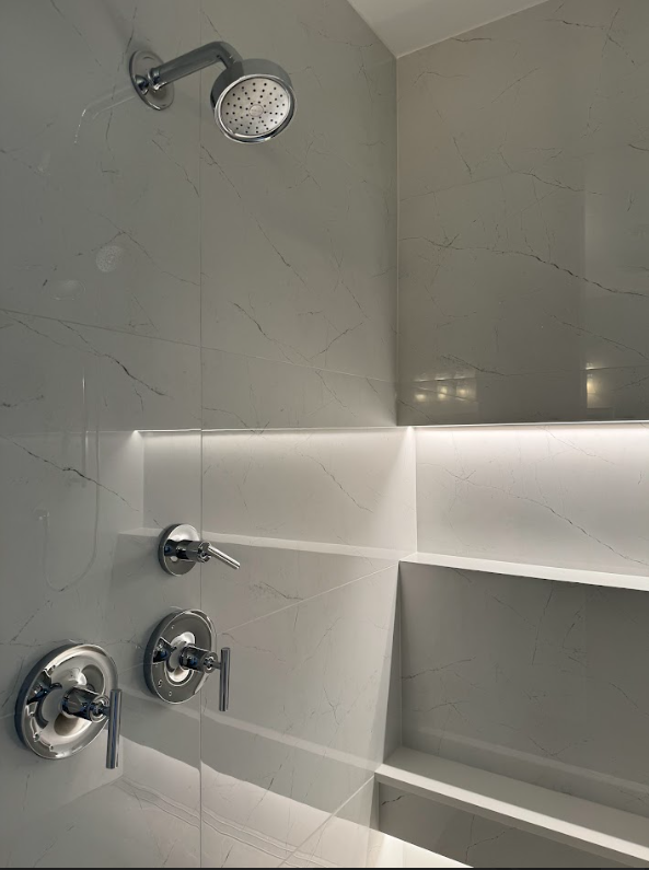 Modern shower with a round showerhead and three control knobs on grey marble wall with built-in shelves, illuminated by white LED lighting.