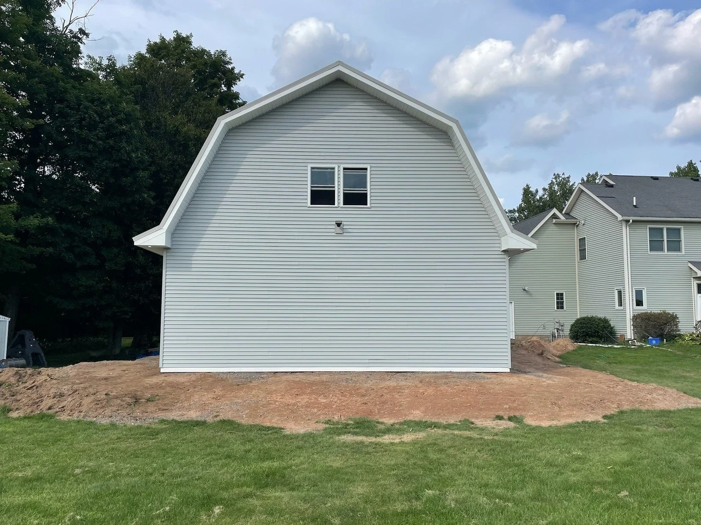 Side view of a house under construction with a large unfinished dirt area in the backyard and neighboring houses in the background.