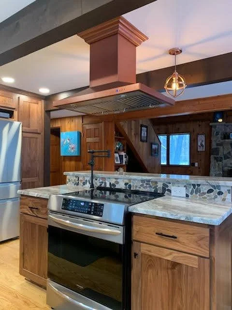 Kitchen with wooden cabinets, granite countertops, a stainless steel oven, and a copper range hood. There is a mosaic tile backsplash, a metal pendant light, and a window in the background.