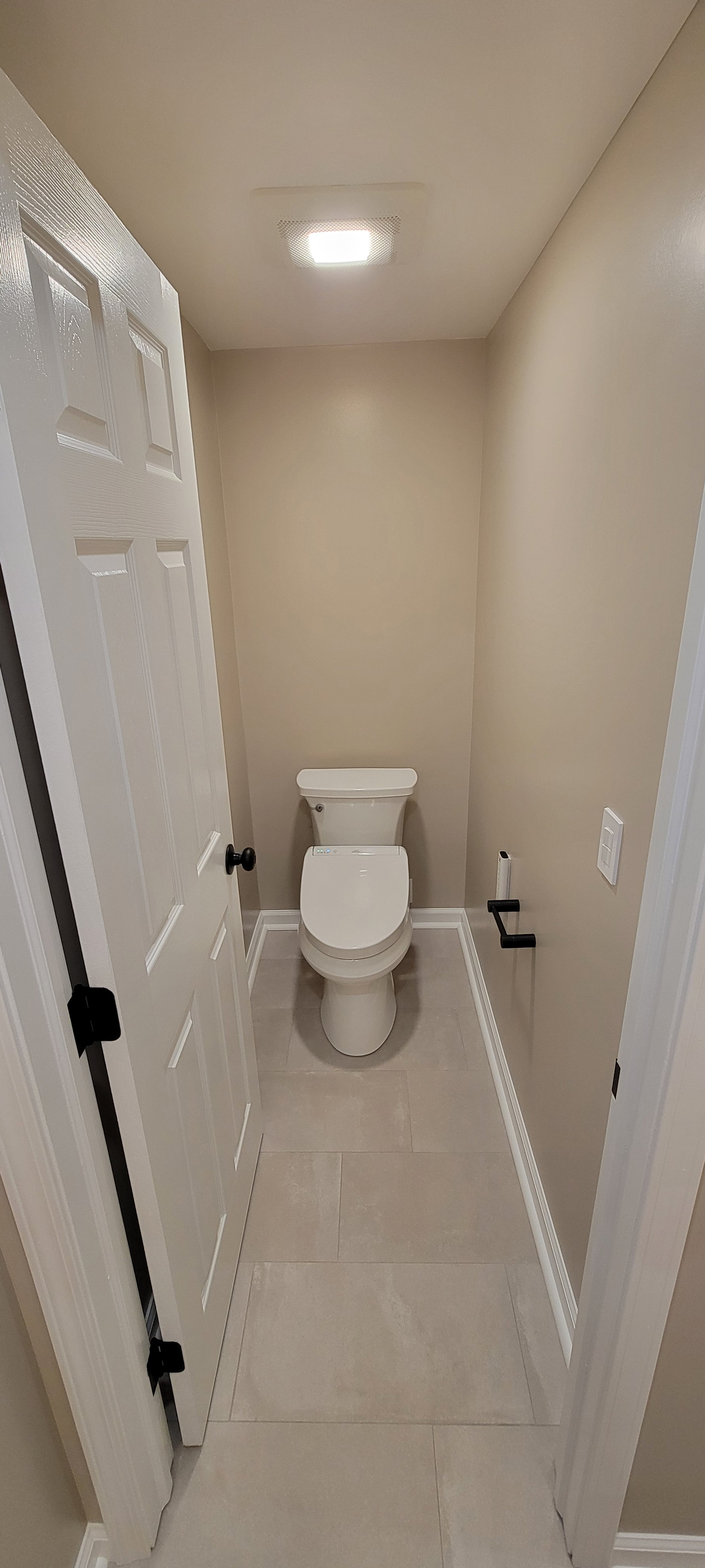 A small, beige-colored bathroom with a white toilet and a closed white door. The floor is tiled, and there is a black toilet paper holder and a light switch on the wall.