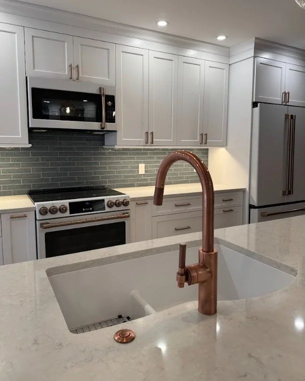 Modern kitchen with white cabinets, gray brick backsplash, stainless steel microwave and refrigerator, and a marble countertop with a copper faucet over a sink.