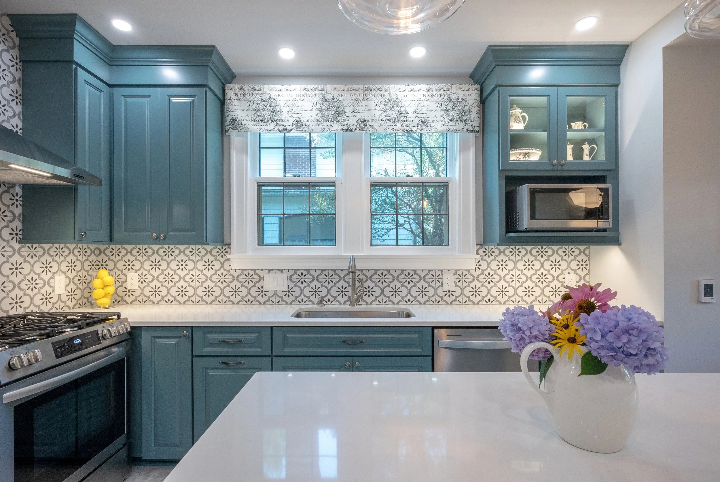 Kitchen with blue cabinets, patterned backsplash, window above sink, flowers in a white vase on the white countertop.