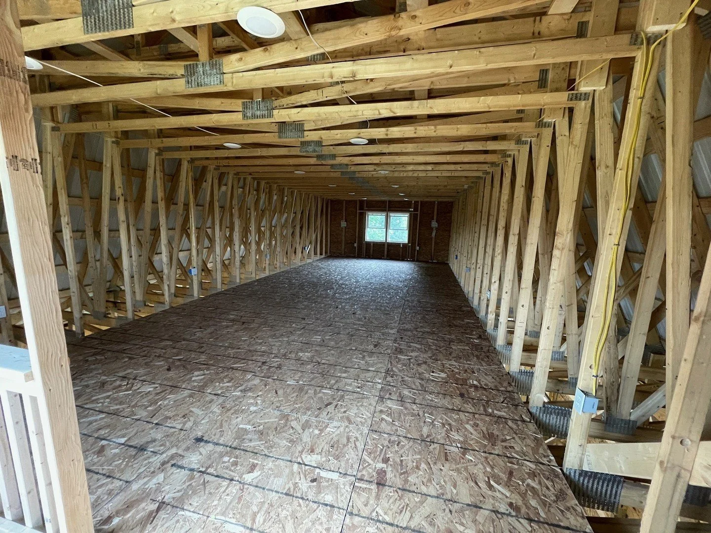 The interior of a building under construction with wooden framing, exposed beams, and a plywood floor, with a small window in the back wall and electrical wiring visible along the walls and ceiling.