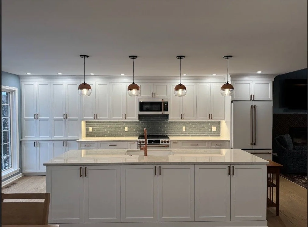 Modern white kitchen with an island, greenish-gray subway tile backsplash, stainless steel appliances, pendant lights, and a large window.