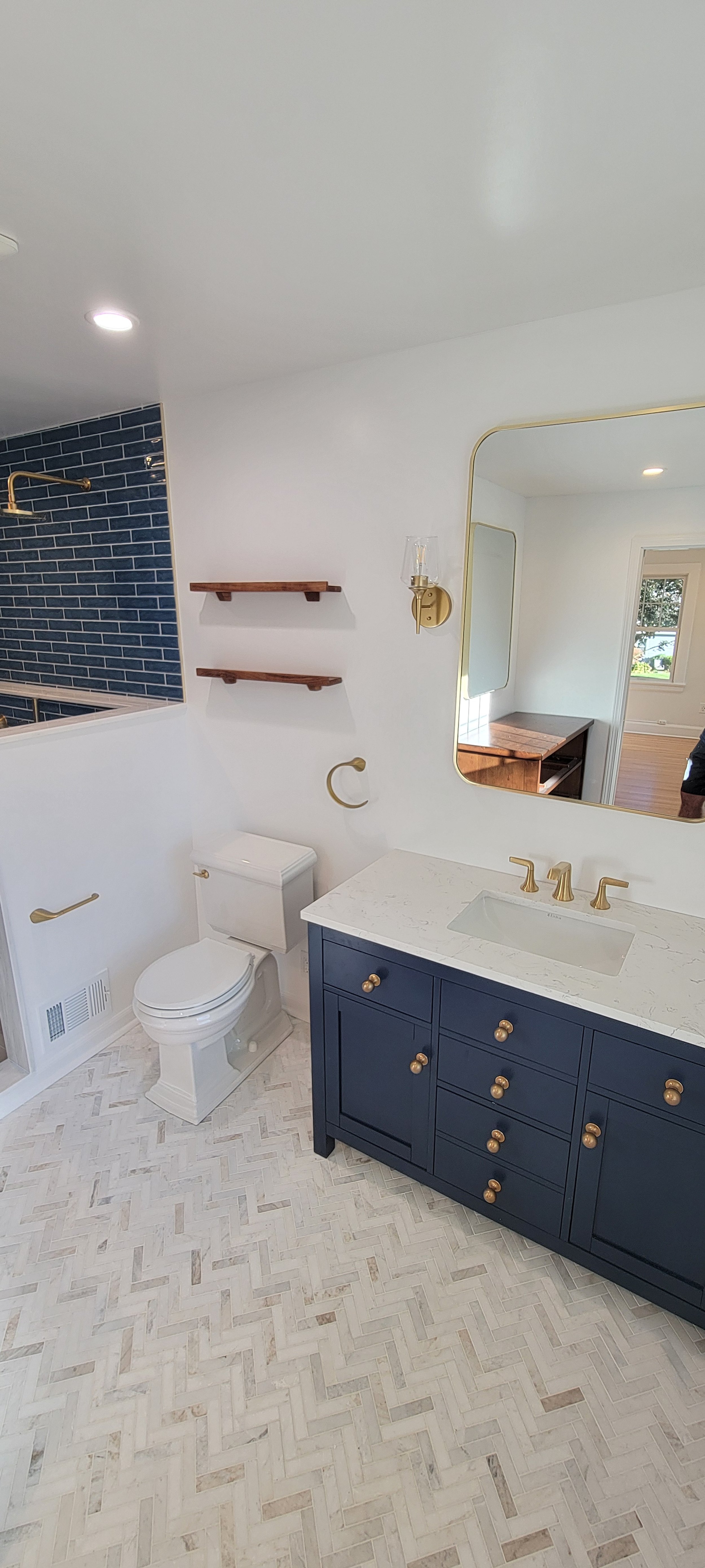 A modern bathroom with white walls, a navy blue vanity with gold knobs, and a marble countertop. There is a large mirror above the vanity, gold fixtures, a toilet, and two wooden shelves on the wall. The bathroom floor has a herringbone pattern with 