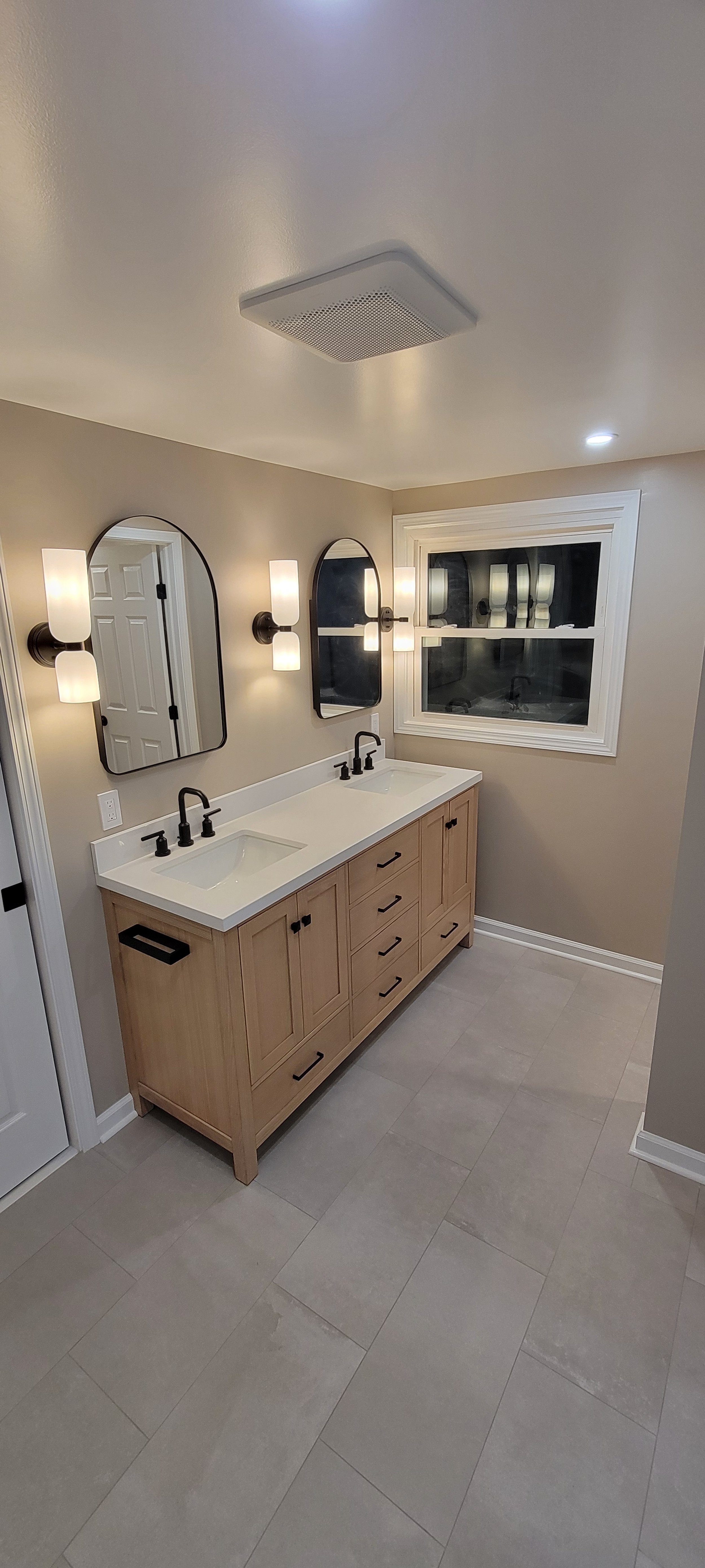A modern bathroom with a double vanity, two black sinks, black faucets, two oval mirrors, wall-mounted light fixtures, a window, beige walls, and light-colored tile flooring.