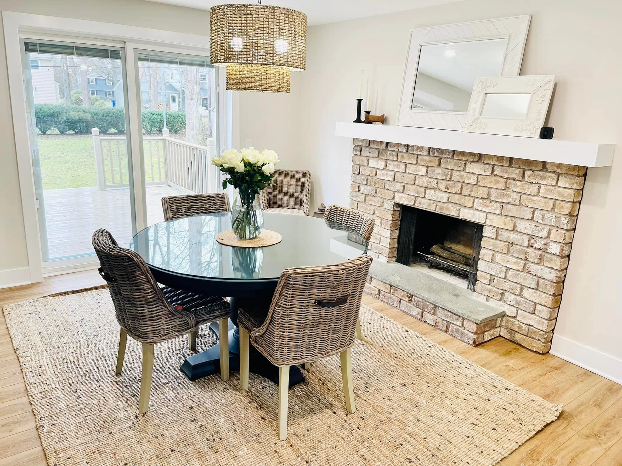 Dining room with round glass table and six wicker chairs, a vase of white roses on the table, a brick fireplace with two mirrors on the white mantel, and a sliding glass door leading to a backyard deck.