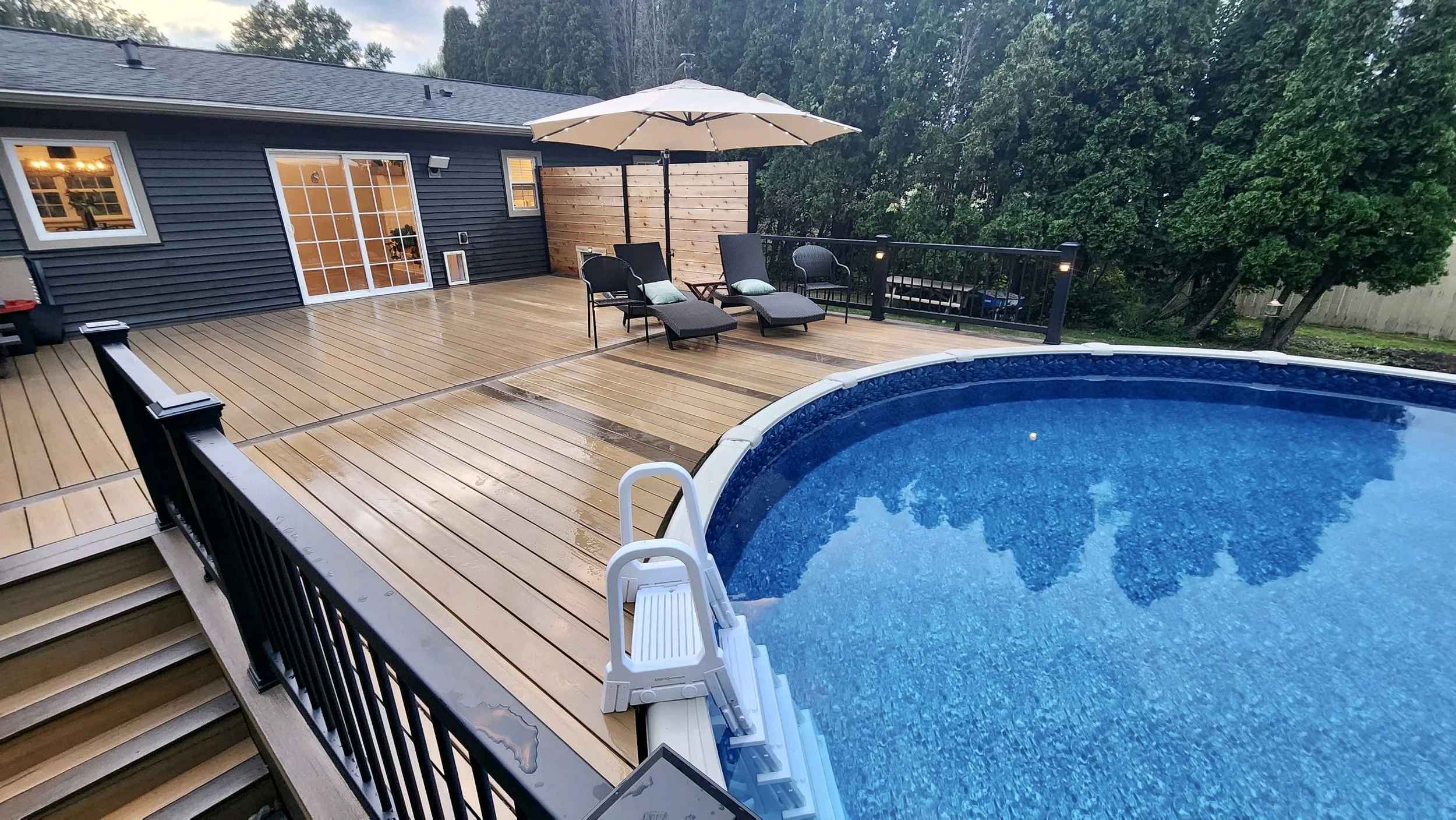 A backyard deck with a swimming pool, lounge chairs with cushions, a large umbrella, and outdoor furniture next to a house with sliding glass doors and windows. There are rain droplets on the deck, indicating recent rain.