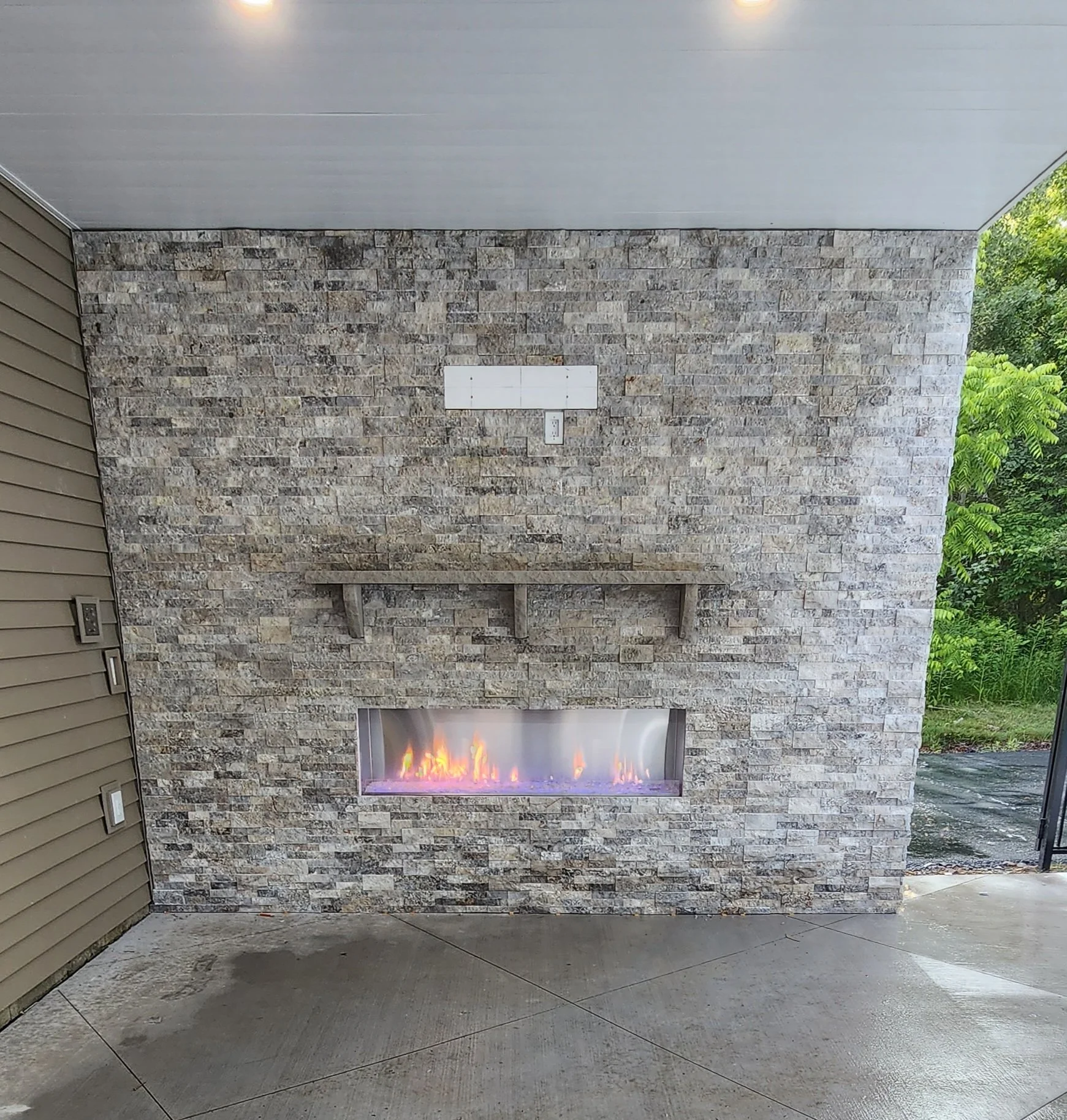 Outdoor patio wall with a rectangular gas fireplace, a stone wall with a wooden shelf, and a brick and greenery background.