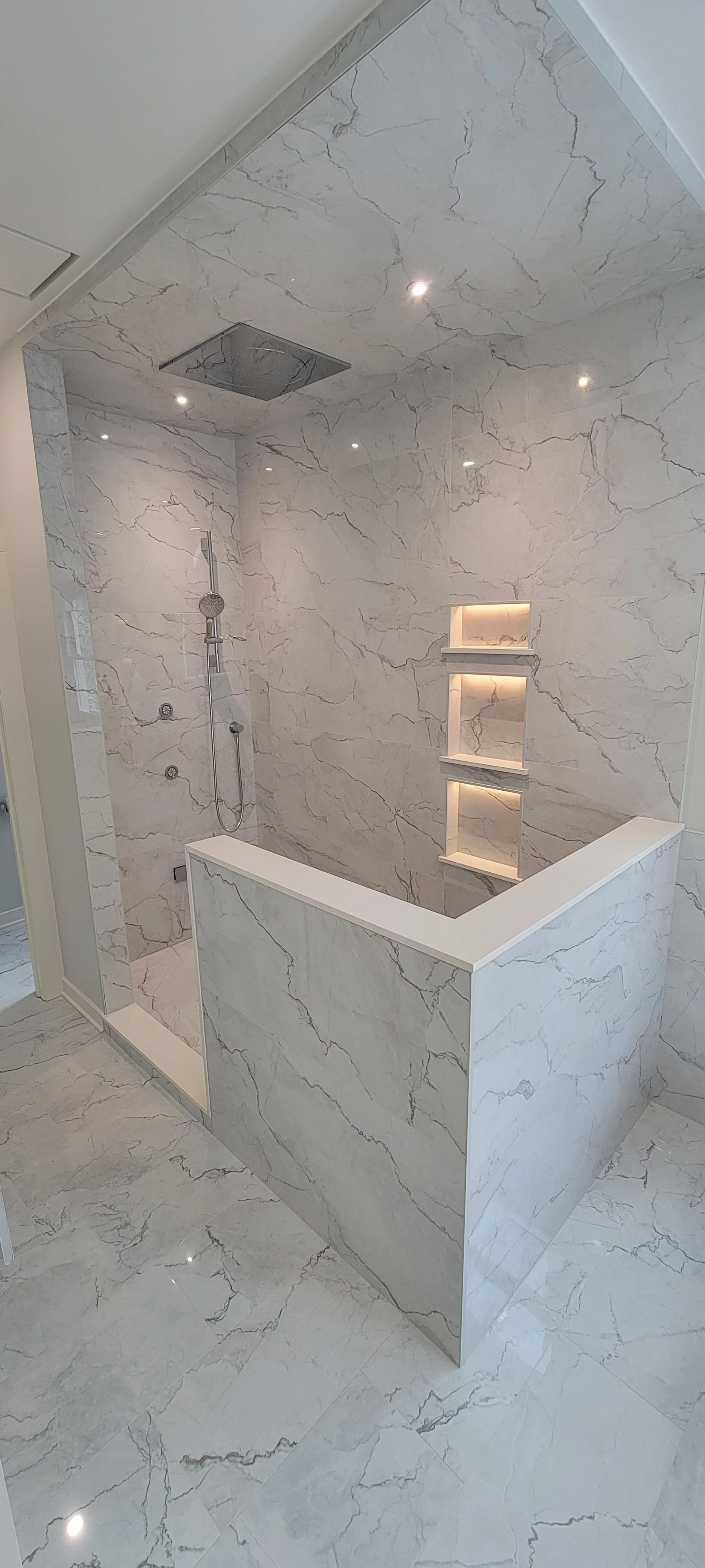 Modern bathroom with marble tiles on floor, walls, and shower area, featuring built-in shelves with backlighting, a ceiling-mounted showerhead, and a partial wall around the shower.