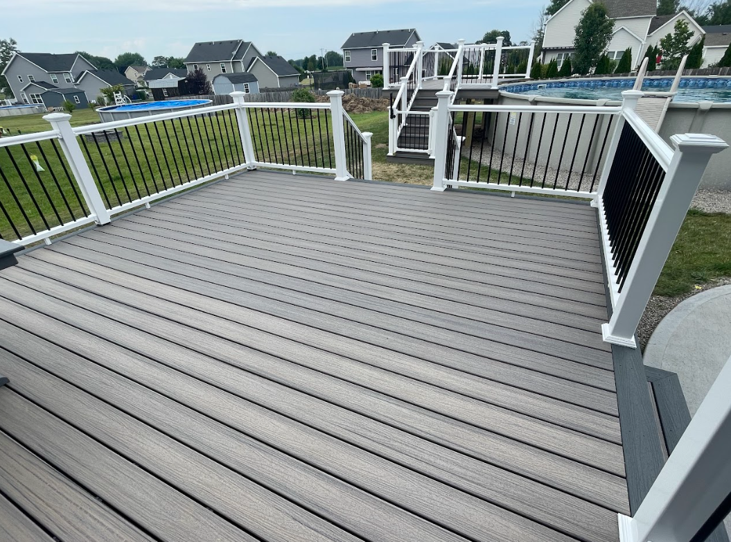 A rooftop deck with gray wood planks, white and black railing, stairs leading down, and an above-ground pool visible in the backyard of a suburban neighborhood.