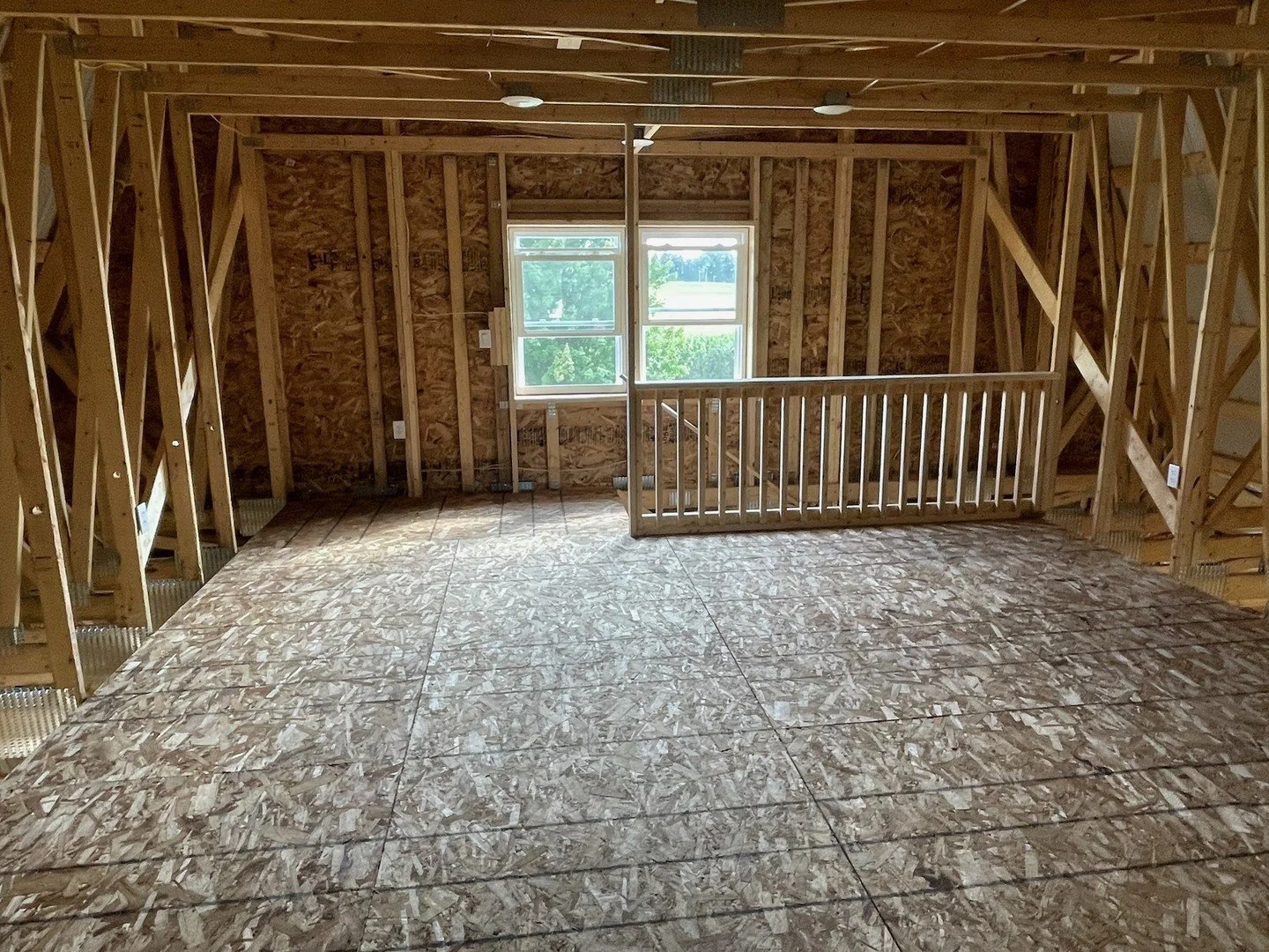 Interior view of an unfinished attic or upper floor with exposed wooden framing, OSB floor, a window, and a small railing.