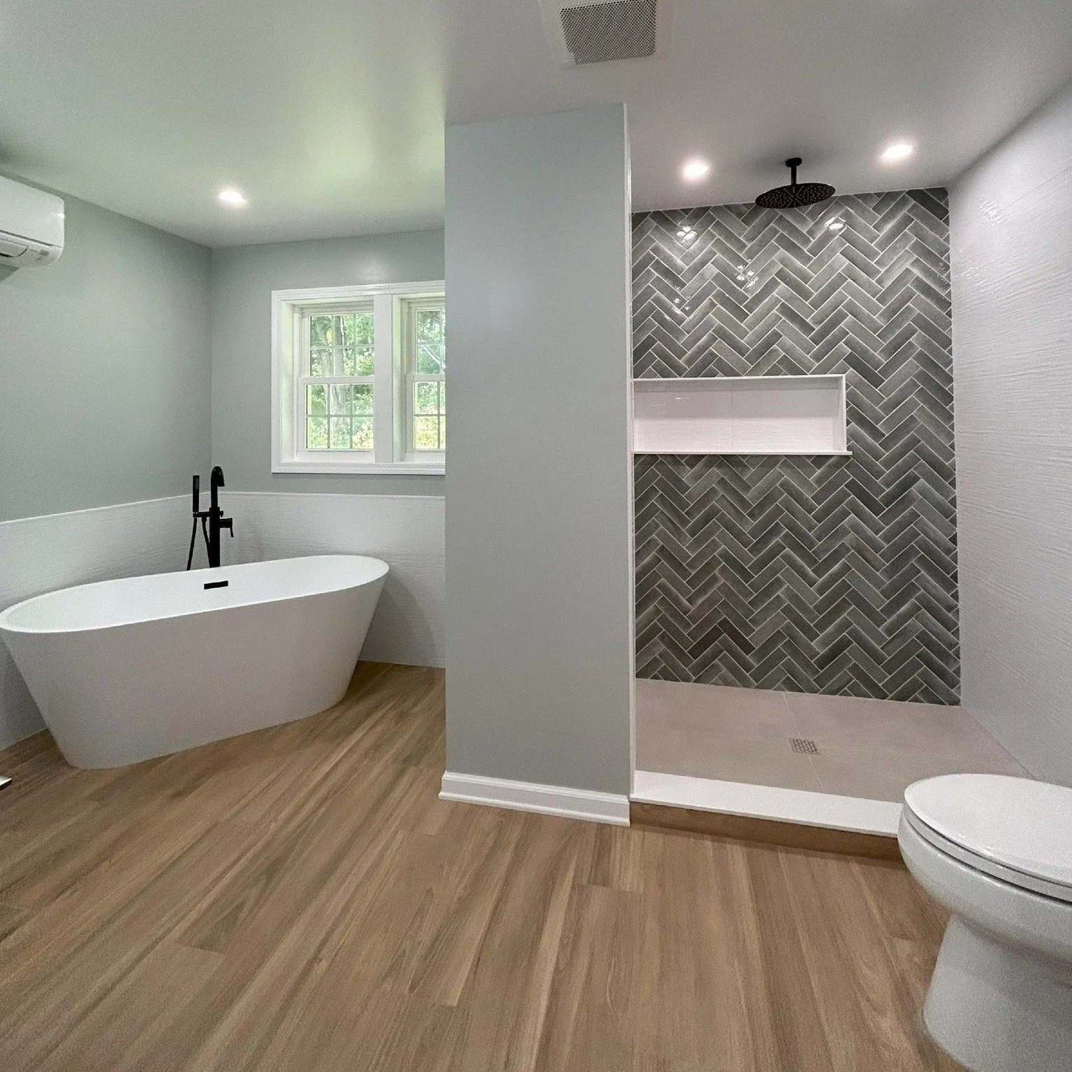 Bathroom with a freestanding bathtub, a window, a walk-in shower with dark gray herringbone tiles and a rain shower head, a toilet, and wood-look flooring.