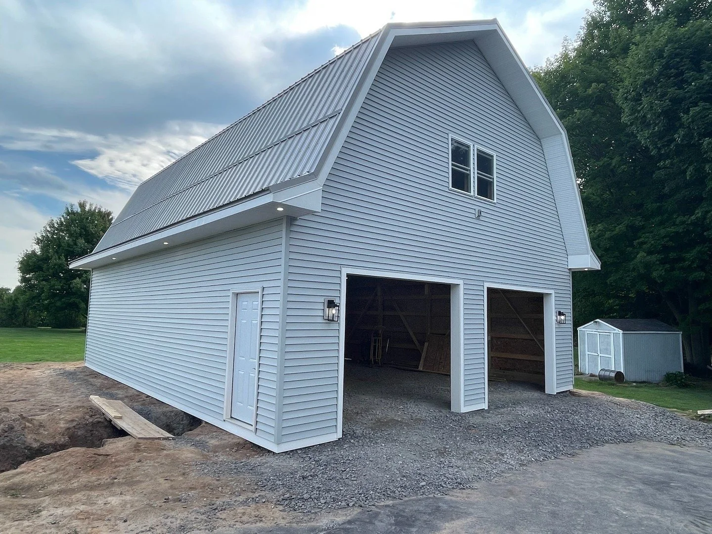 White barn-style garage with two open doors, one side door, and a small window on the upper level, set on a gravel driveway with a small shed and trees in the background.