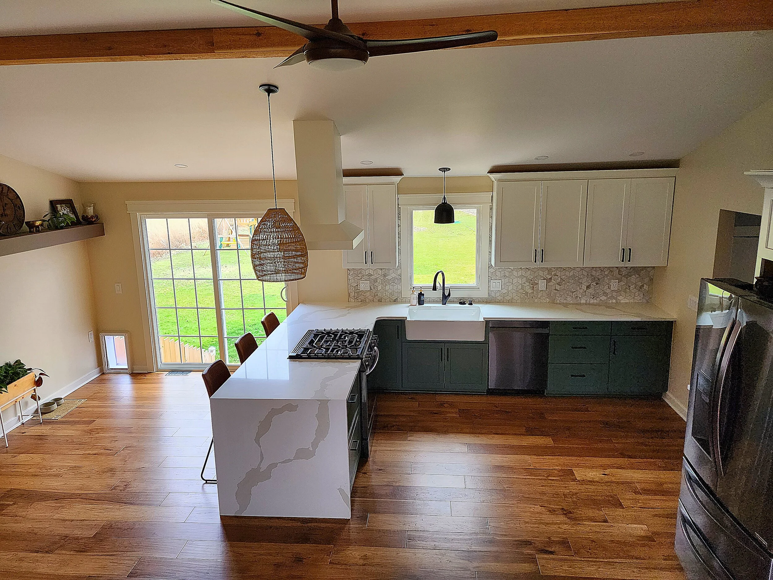 Kitchen with white upper cabinets, dark lower cabinets, marble island, and hardwood floors. Large window above sink, sliding glass door leading outside, pendant lights, and ceiling fan visible.