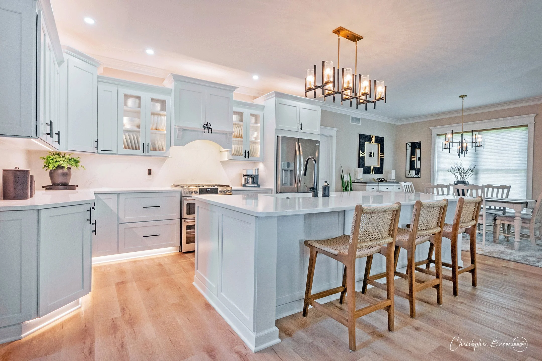 Bright, modern kitchen with white cabinets, a large central island with a white countertop, four wooden bar stools, stainless steel appliances, light hardwood flooring, and contemporary lighting fixtures.