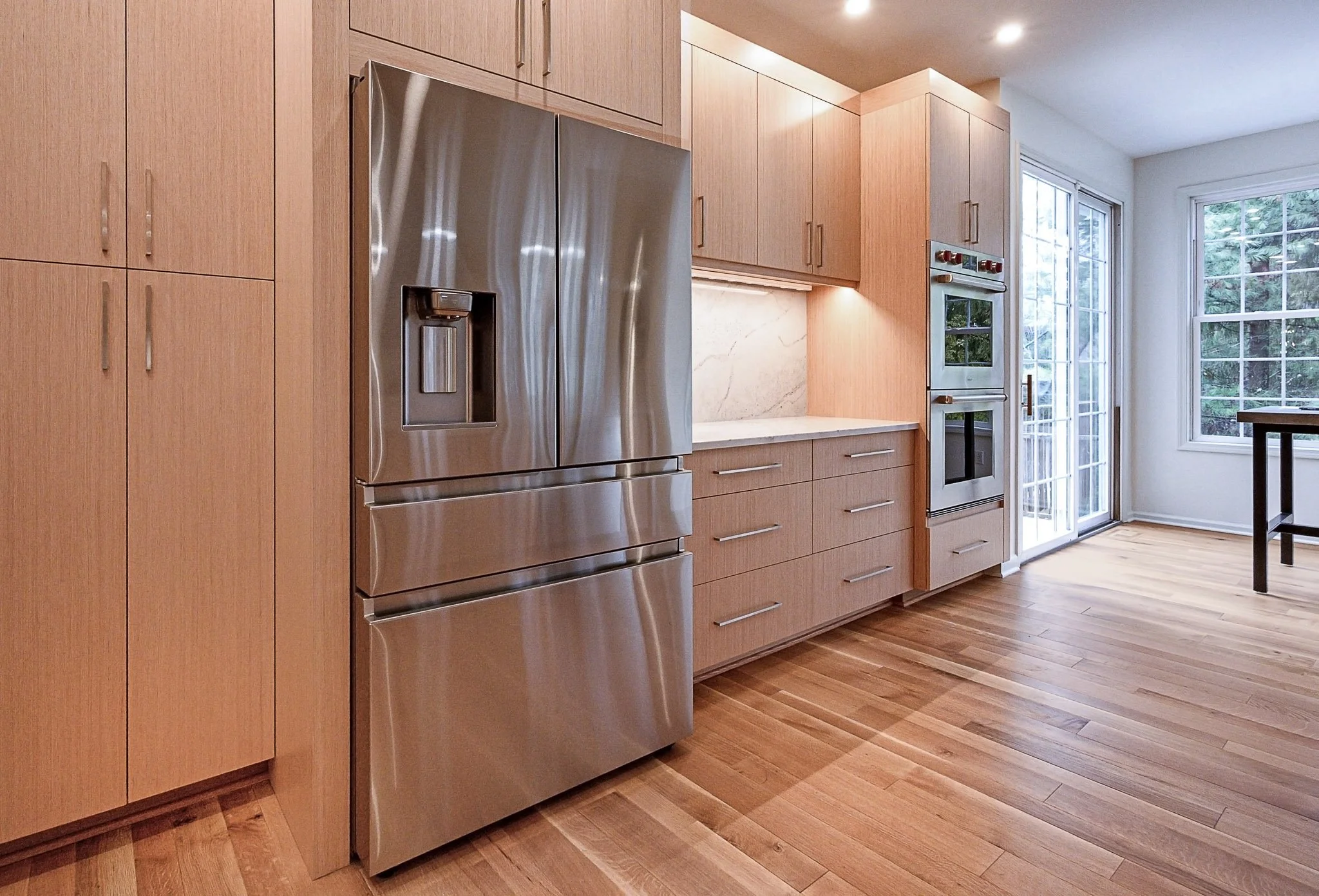 Modern kitchen with wooden cabinets, stainless steel refrigerator, built-in oven, and hardwood flooring, adjacent to large windows and a sliding glass door.