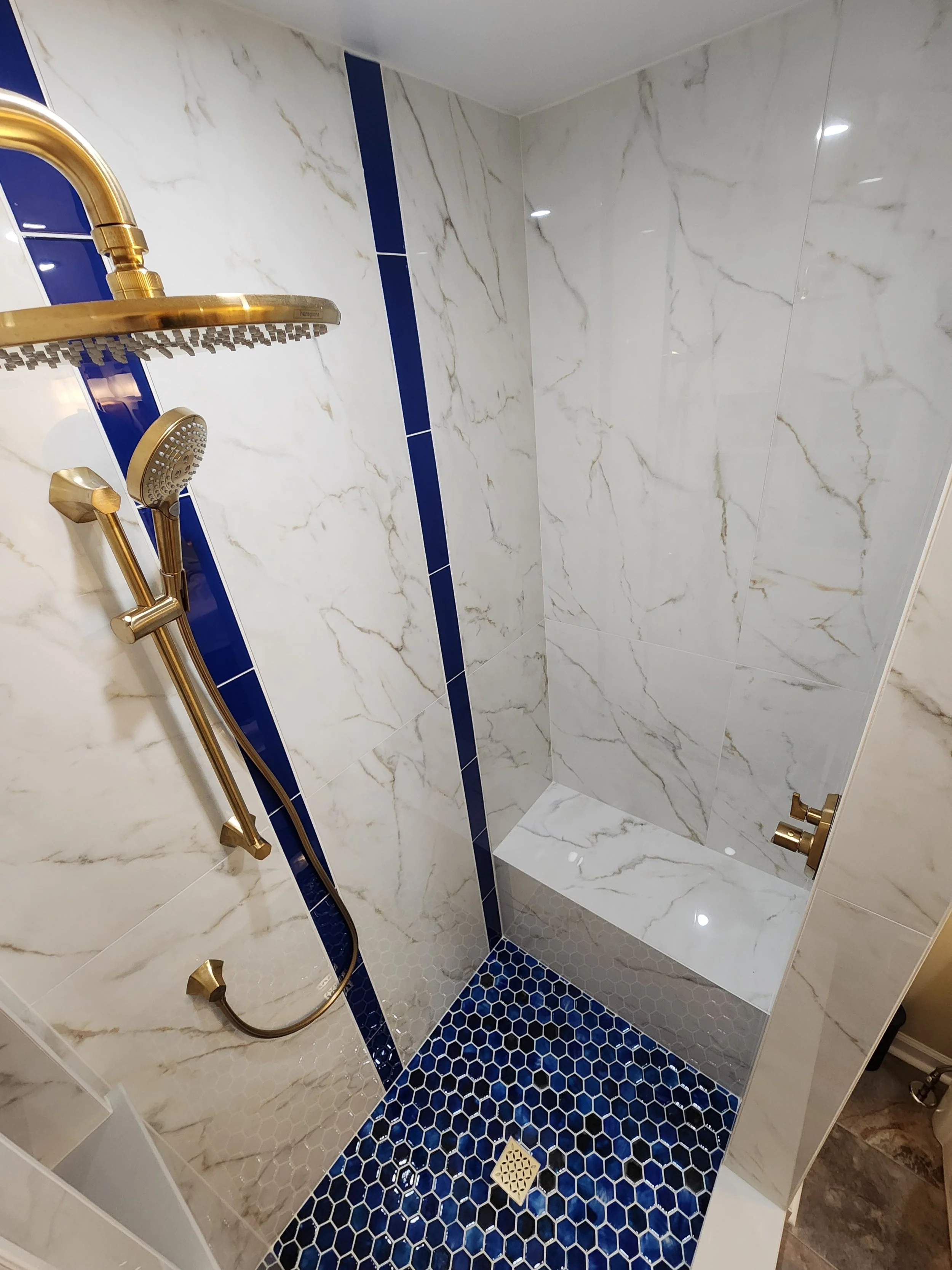 Shower area with white marble walls, blue tile accents, a blue hexagon tile floor, a built-in bench, and a gold showerhead and fixtures.