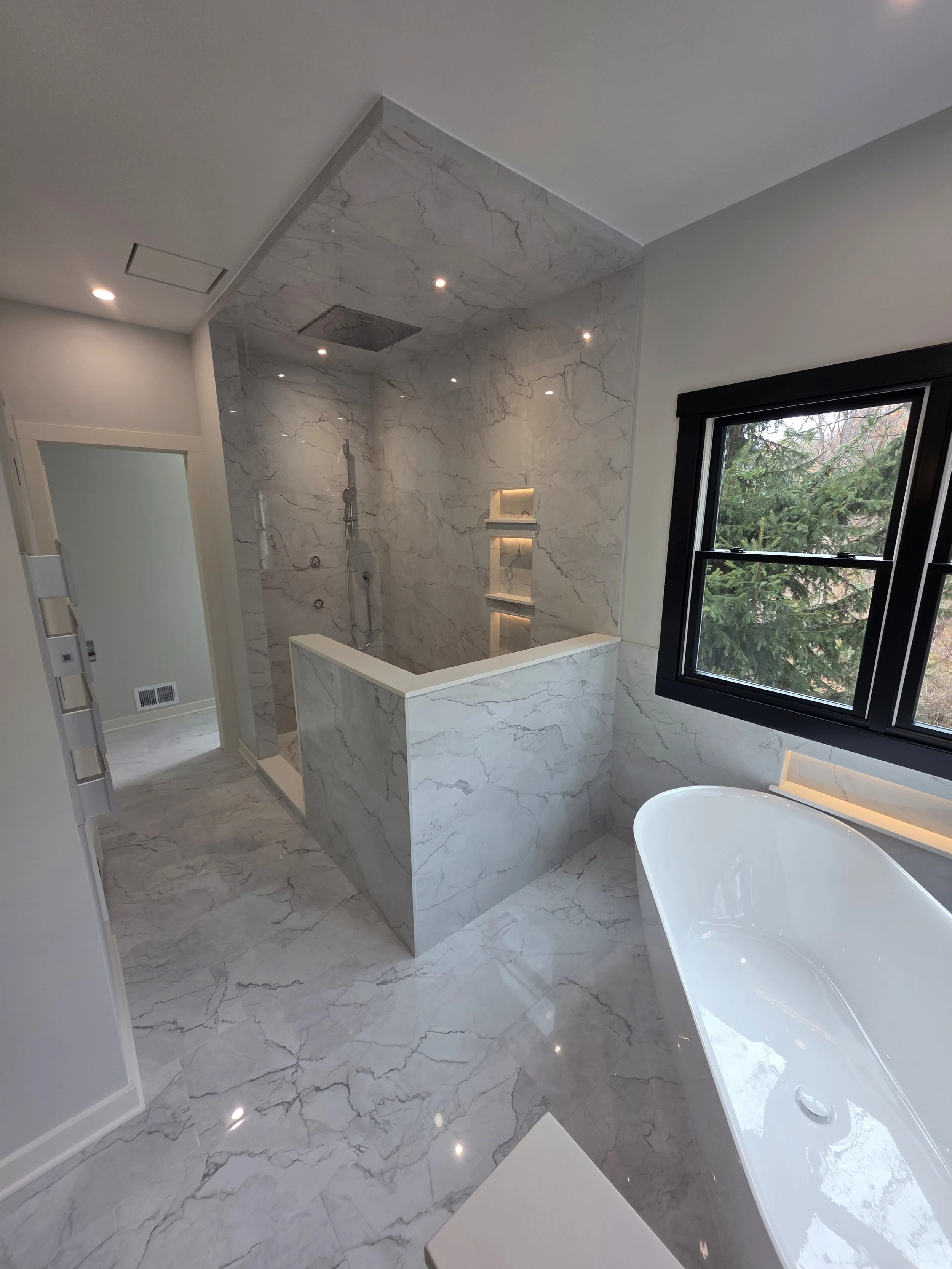 Luxury bathroom with marble tiles, walk-in shower with niches, freestanding bathtub, and large window showing trees outside.