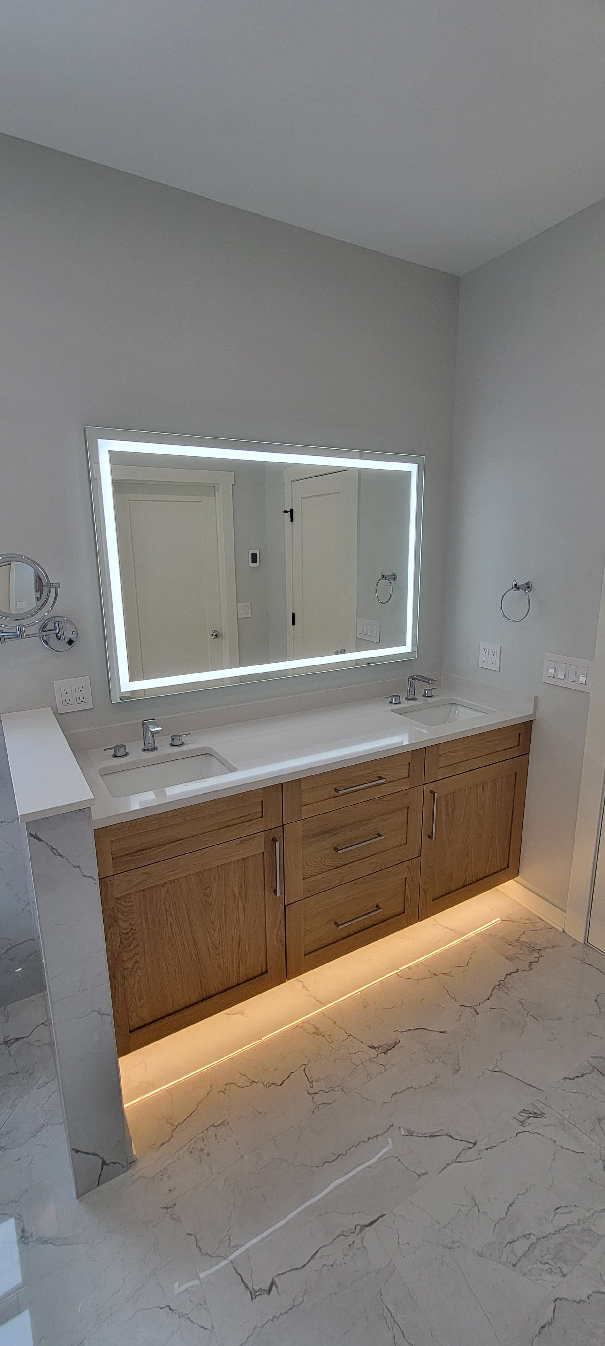 Modern bathroom vanity with a large illuminated mirror, dual sinks, wooden cabinet, marble floor, and soft under-cabinet lighting.
