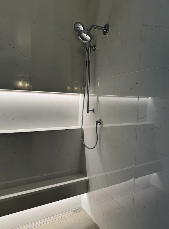 A modern shower with gray marble walls, a handheld showerhead, and built-in white shelves with integrated lighting.