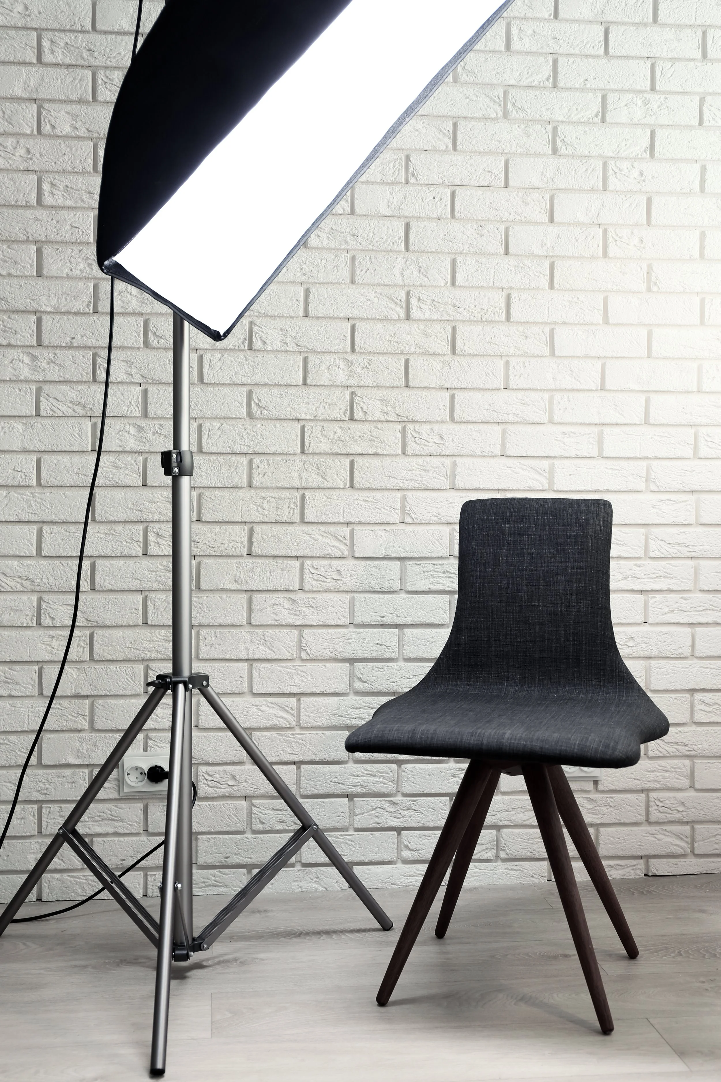 Photo of a photography studio setup with a black chair, large studio light on a stand, white brick wall, and electrical outlet on the floor.