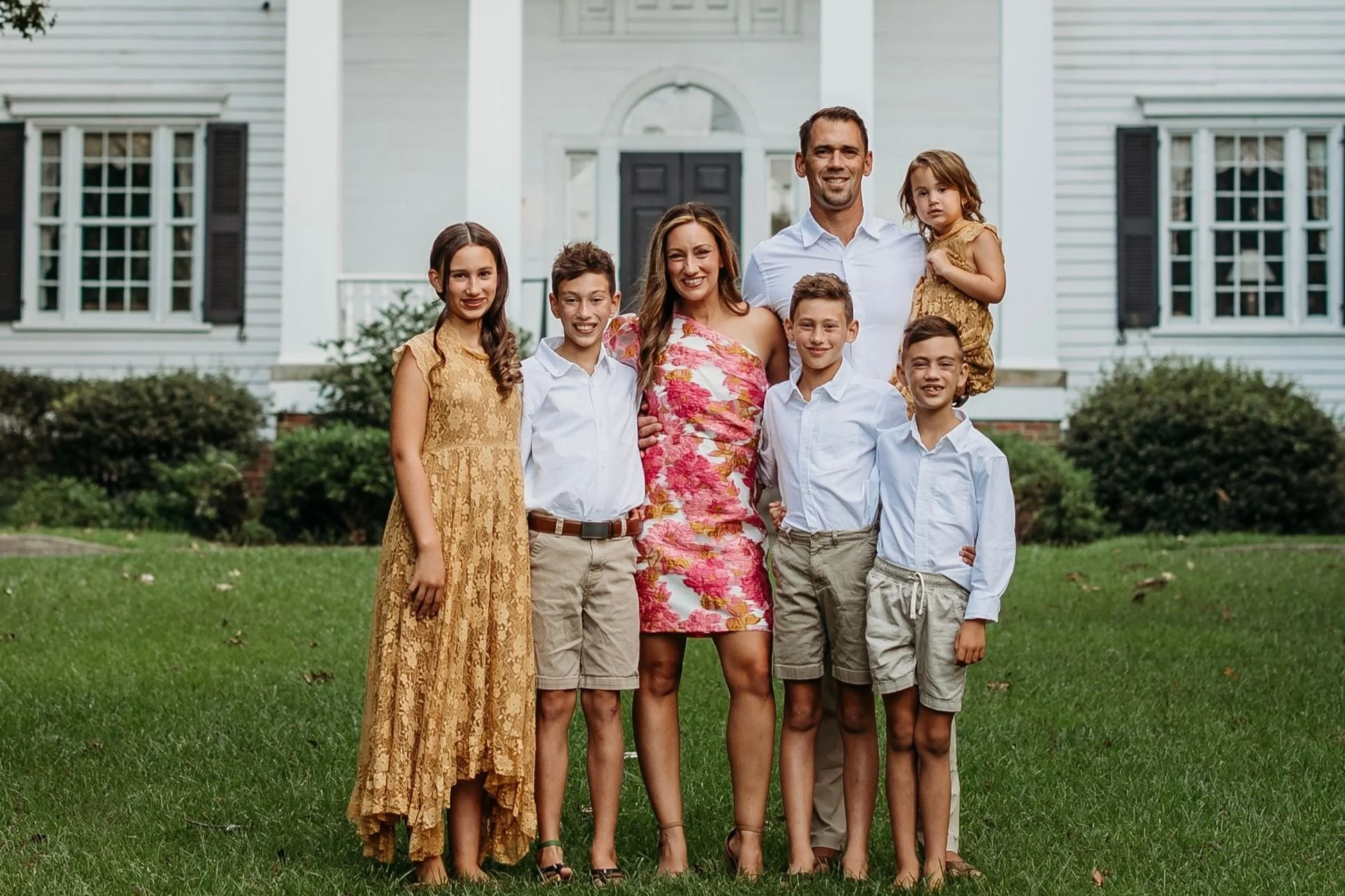 Family of seven standing on grass in front of a white house with black shutters. Four children, a woman, and a man, all smiling and posing for the photo.