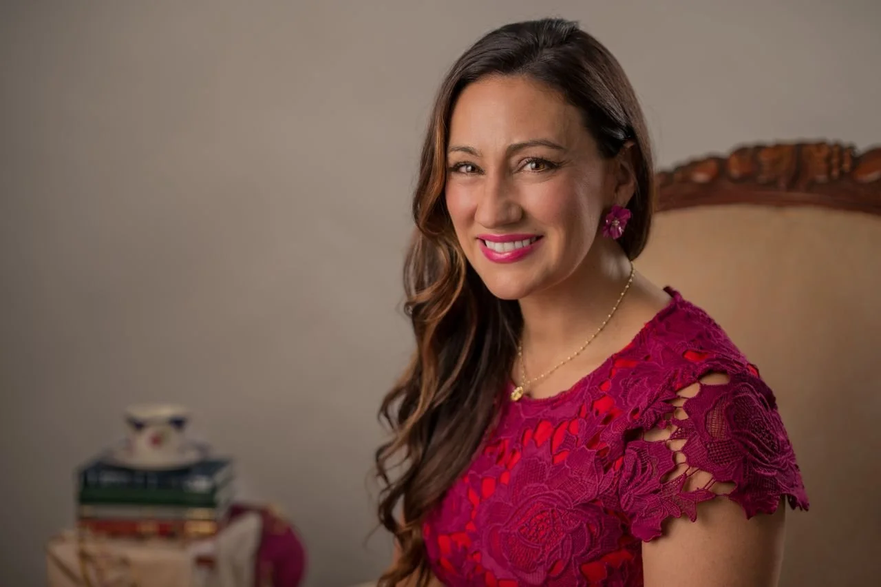 A woman with long wavy brown hair smiling and wearing a pink lace dress, pink earrings, and a gold necklace, sitting on an ornate wooden chair.
