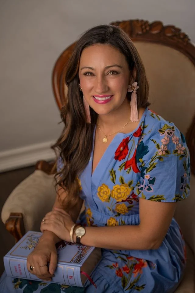 A woman with long brown hair, wearing a floral blue dress and pink tassel earrings, sitting on a vintage wooden chair. She is smiling and holding a book, with a gold necklace and watch, in a well-lit room with neutral walls and a framed mirror in the background.